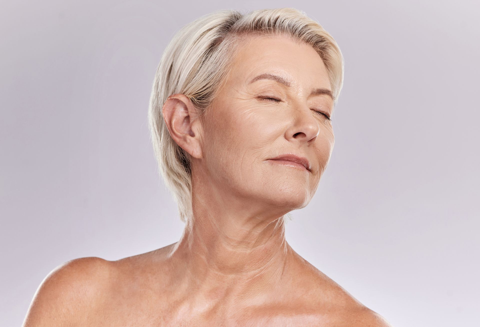 Woman with short blonde hair, eyes closed, smiling, bare shoulders, against a light gray background.