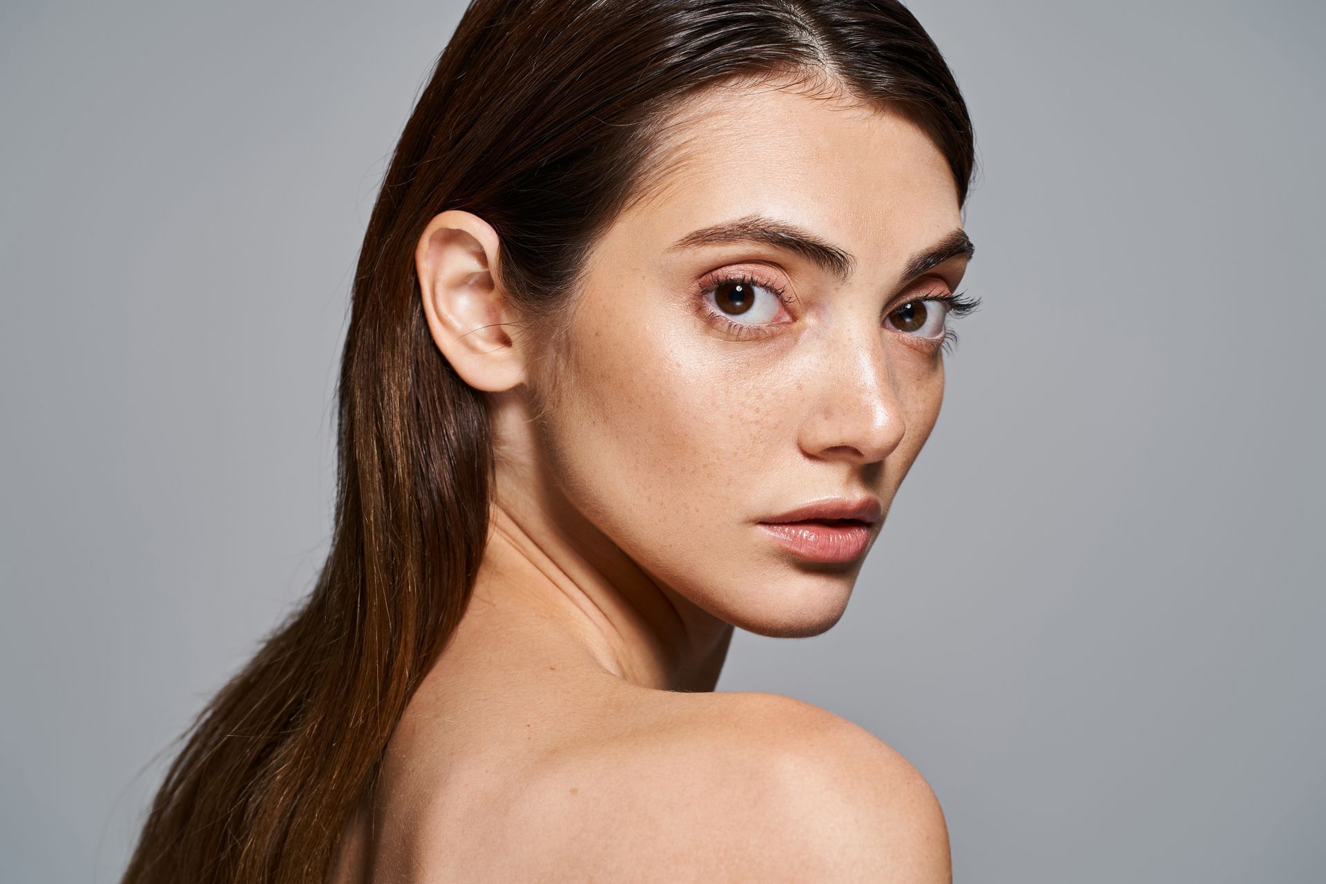 Woman with slicked-back hair looking over shoulder at viewer. Skin tone fair, freckles visible, neutral background.