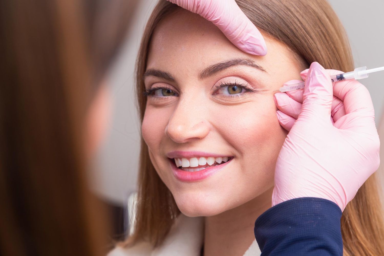 A clinician wearing pink gloves administers a cosmetic injection to a person's eye area in a professional clinic setting.