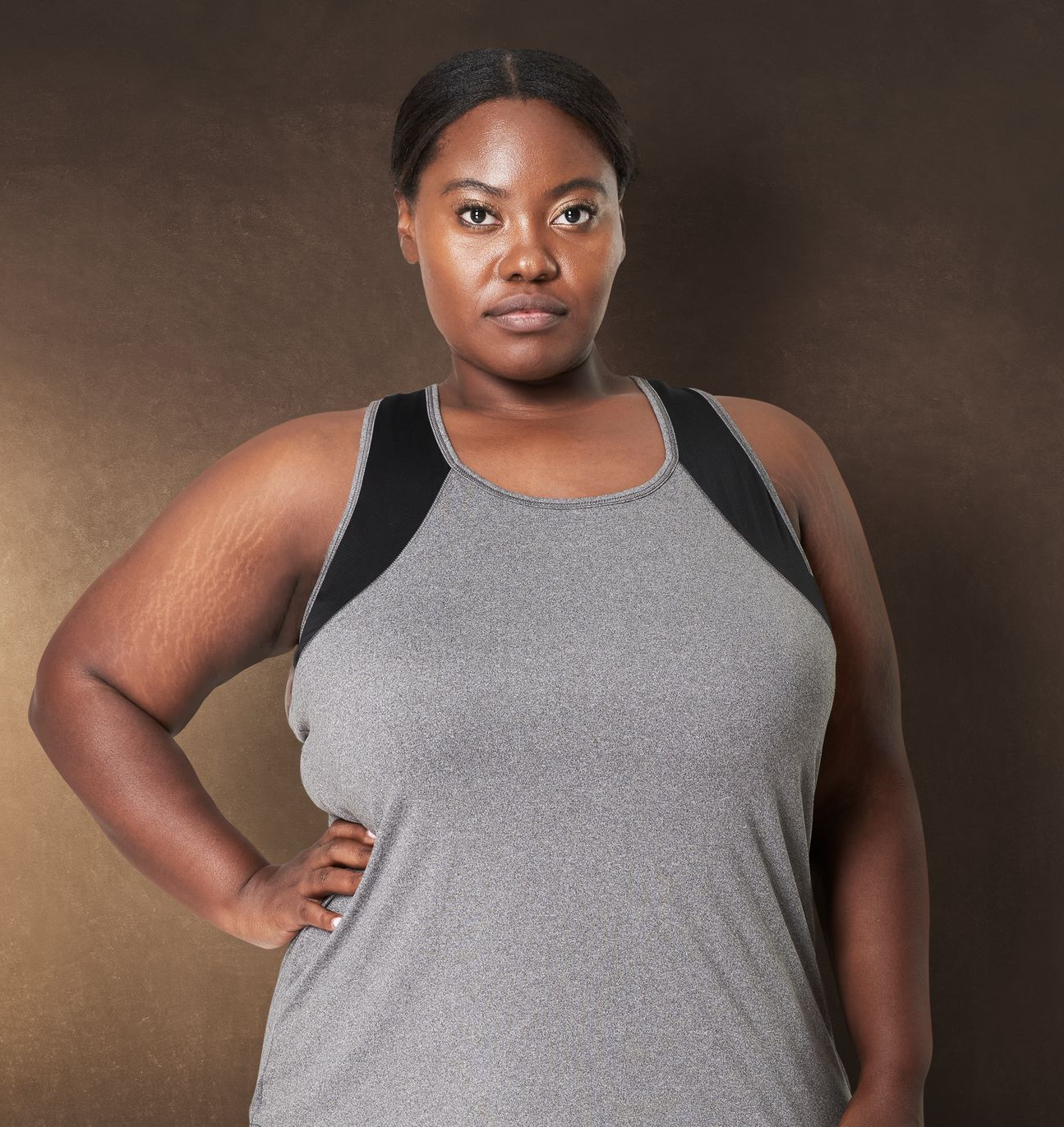 Woman in gray athletic wear, hand on hip, looking at the camera against a brown background.