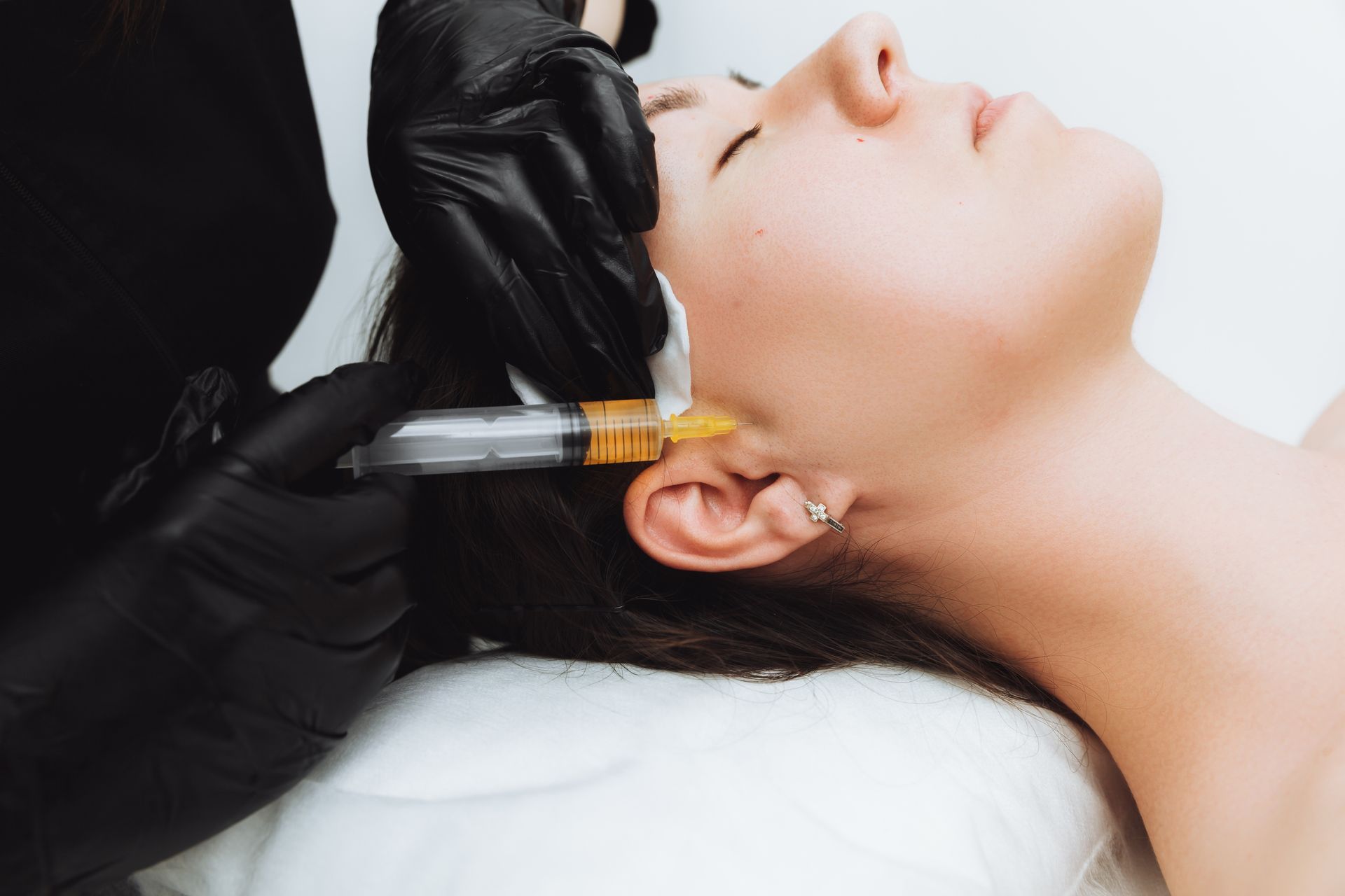 Gloved clinician injecting filler near a woman’s jawline during a cosmetic procedure
