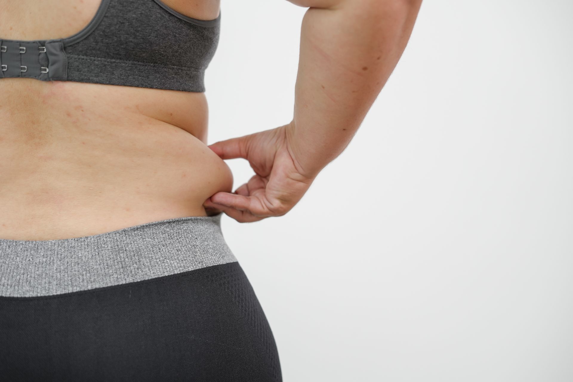 Person pinching side waist in gray sports bra and black leggings against a white background
