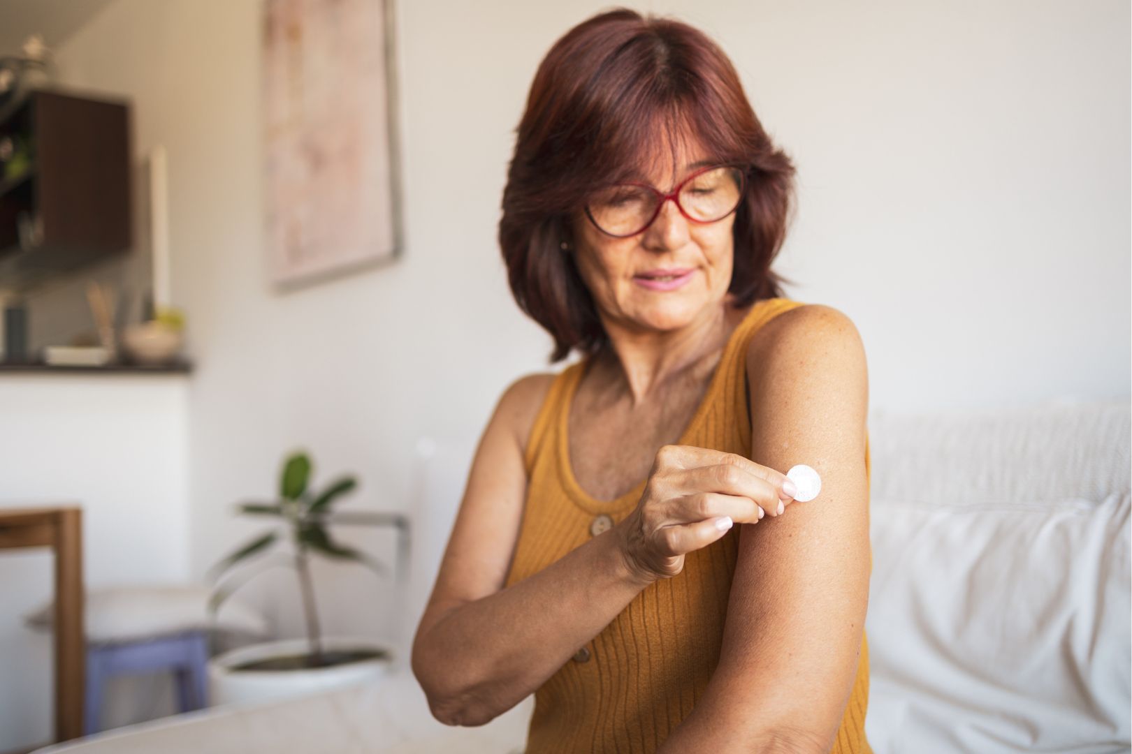 Woman in glasses applying a patch to her upper arm indoors.