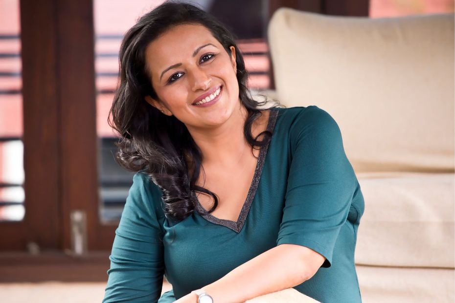 Woman with large, curly brown hair, smiling widely, hands on her cheeks; seated in a white wire chair.