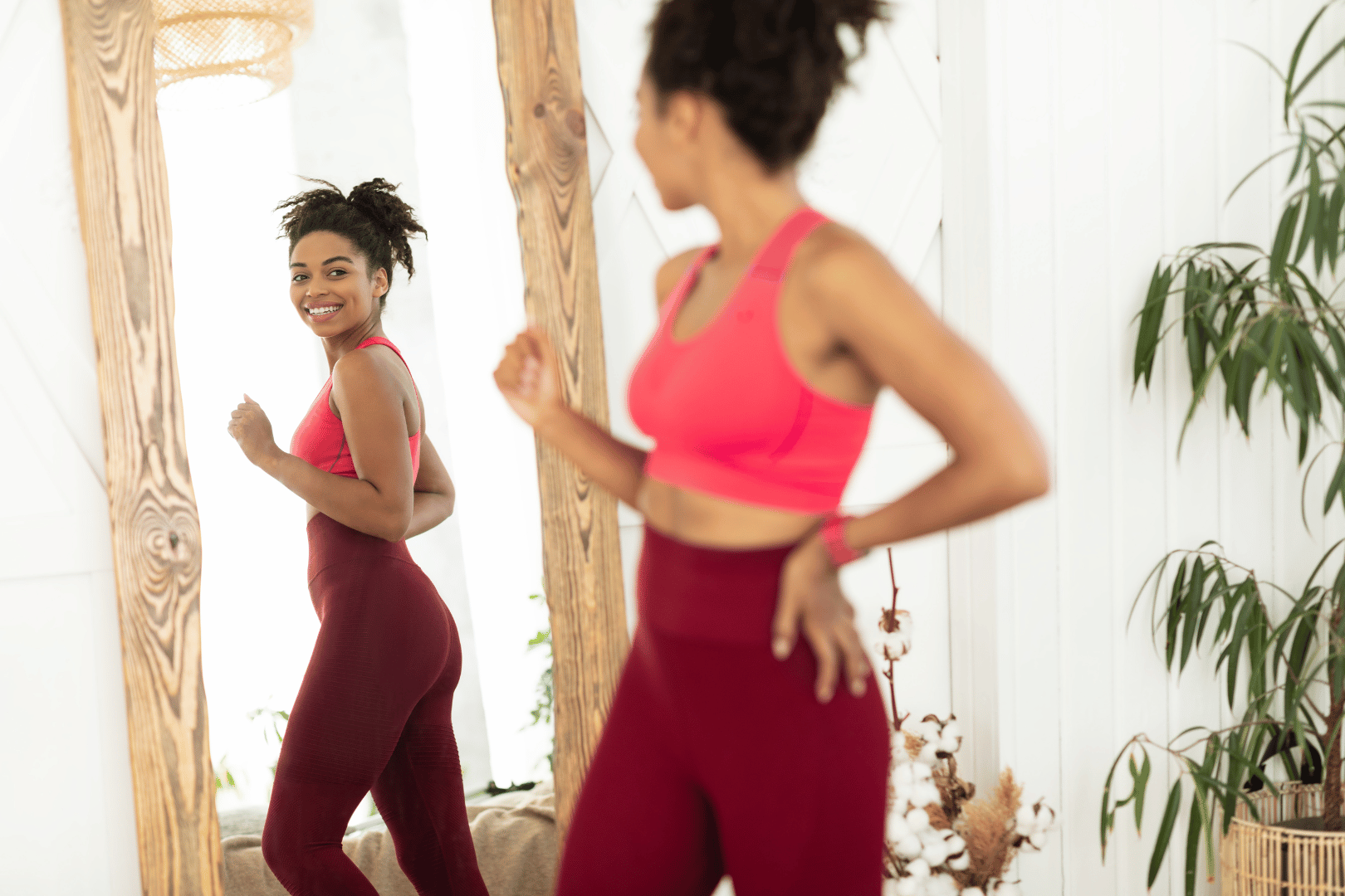 Woman in red workout clothes looks at herself in a mirror, smiling. Bright room, wooden frame.