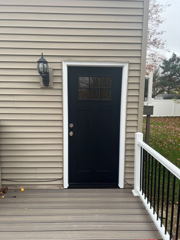 A black door with a glass pane window in a tan siding wall, featuring a wall-mounted light and a white deck railing.