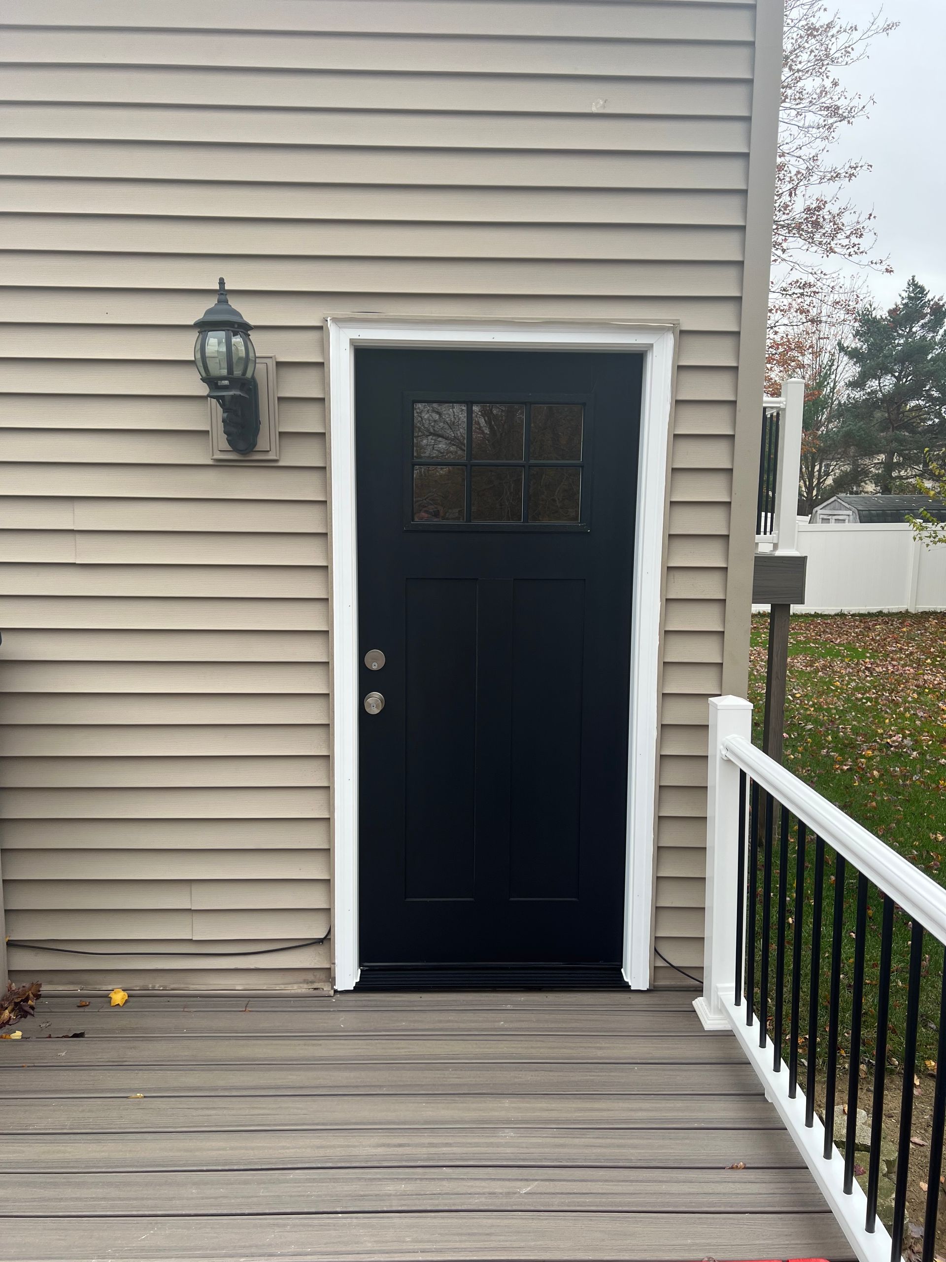 A black exterior door with a window at the top, installed in a tan-sided wall with white trim and a wooden deck nearby.