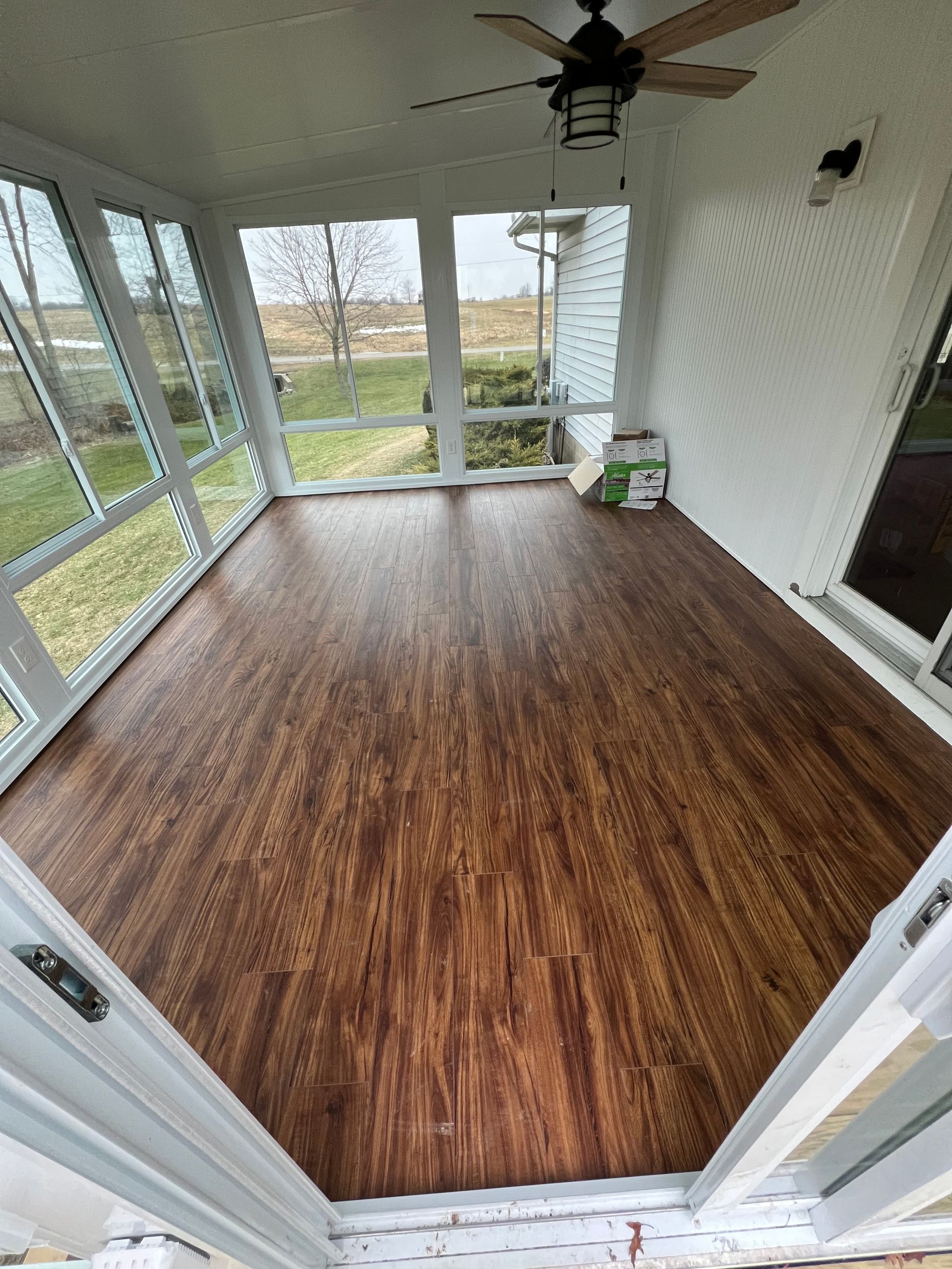 A sunroom with dark wood-look vinyl flooring, large windows, and a ceiling fan, viewed from a doorway.
