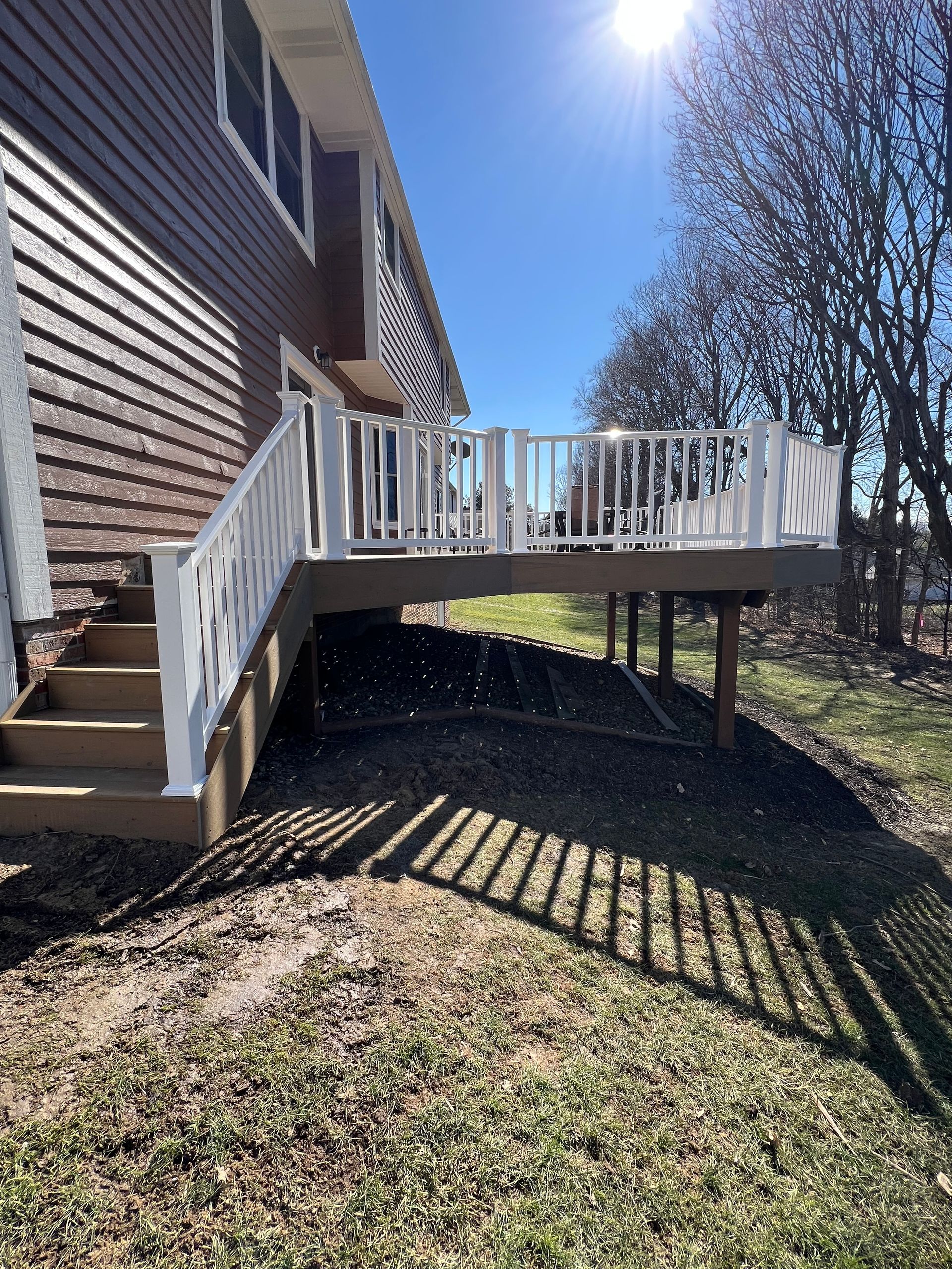A side view of a raised wooden deck with white railings and stairs, attached to the side of a brown-sided house.