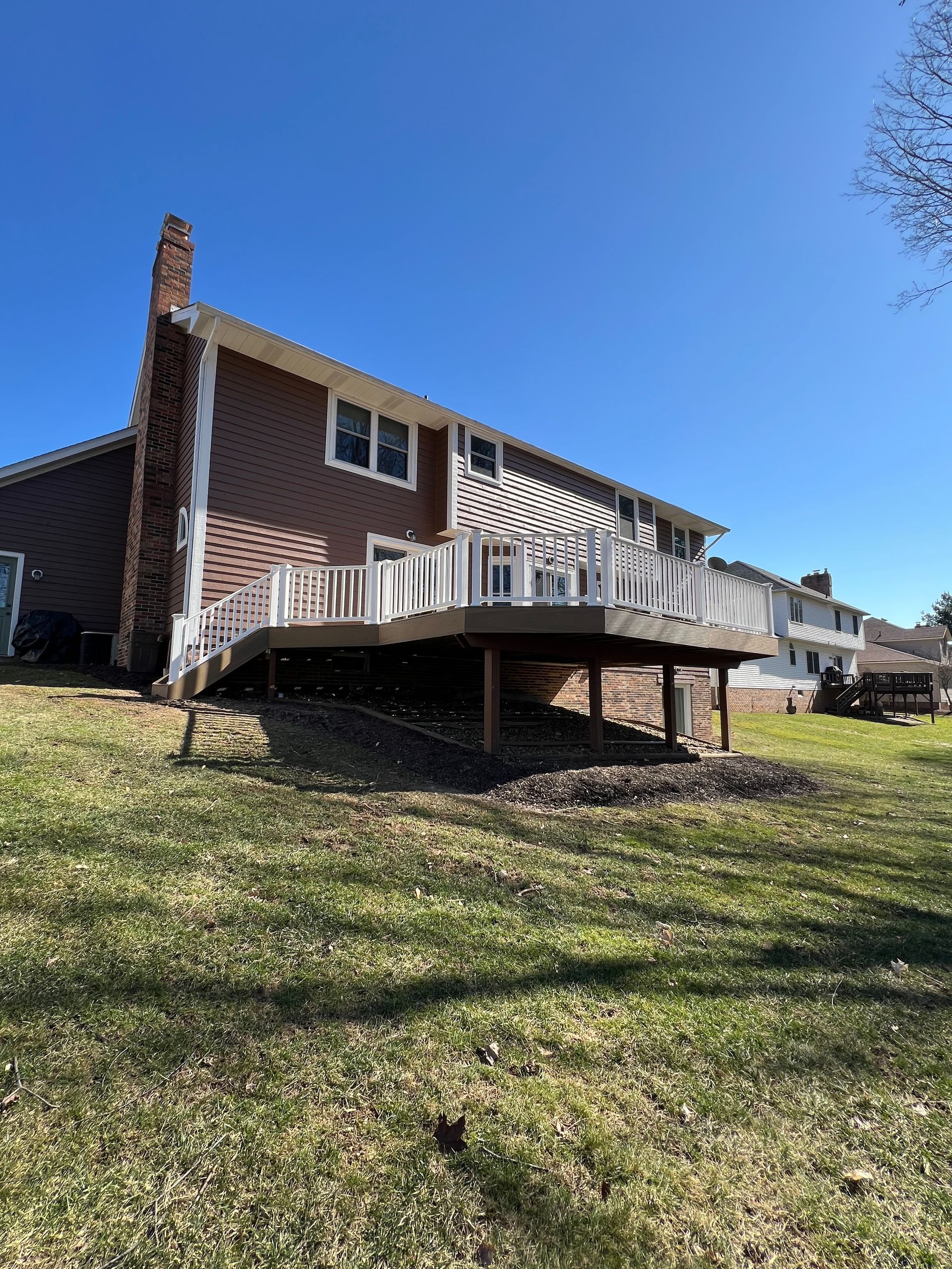 A brown-sided house with a large, elevated light-colored wooden deck in the backyard on a sunny day.