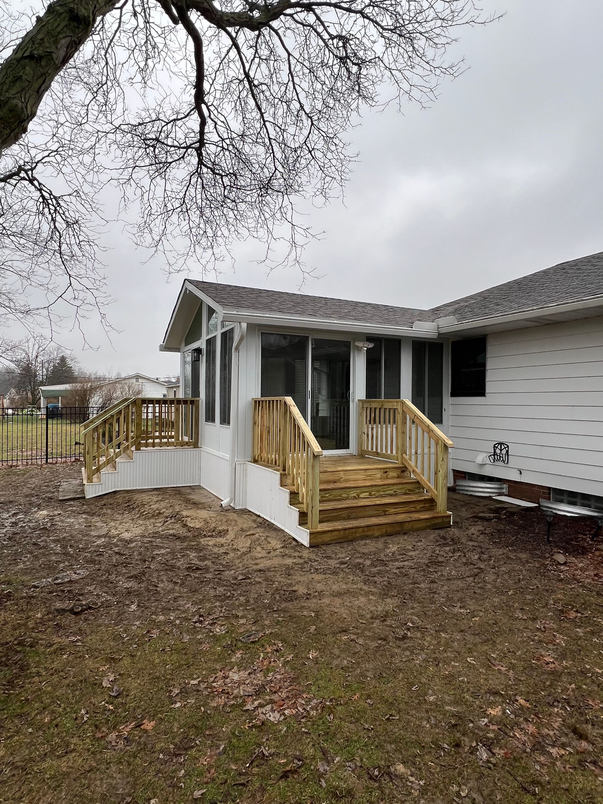 A white house with a sunroom addition, featuring a newly constructed wooden deck and stairs, set in a yard with trees.