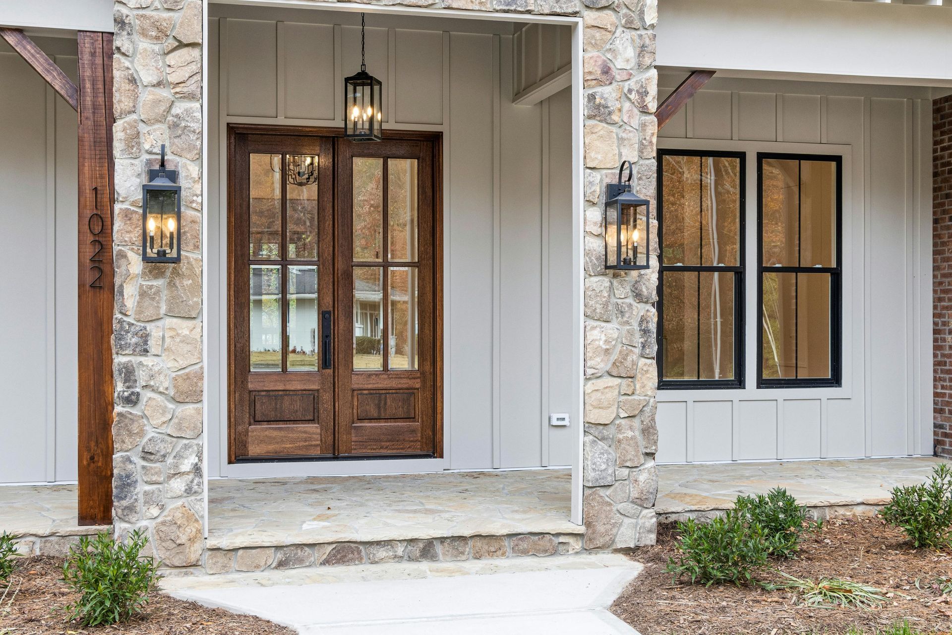 A double-door wooden entry with glass panels, flanked by stone columns and black lanterns on a white home porch.