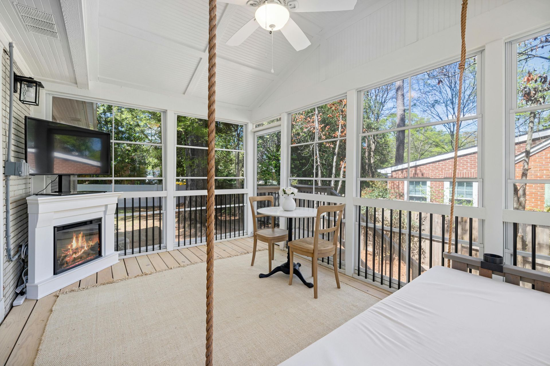 A bright sunroom with a white electric fireplace, a small bistro table with two chairs, and a hanging swing bed.