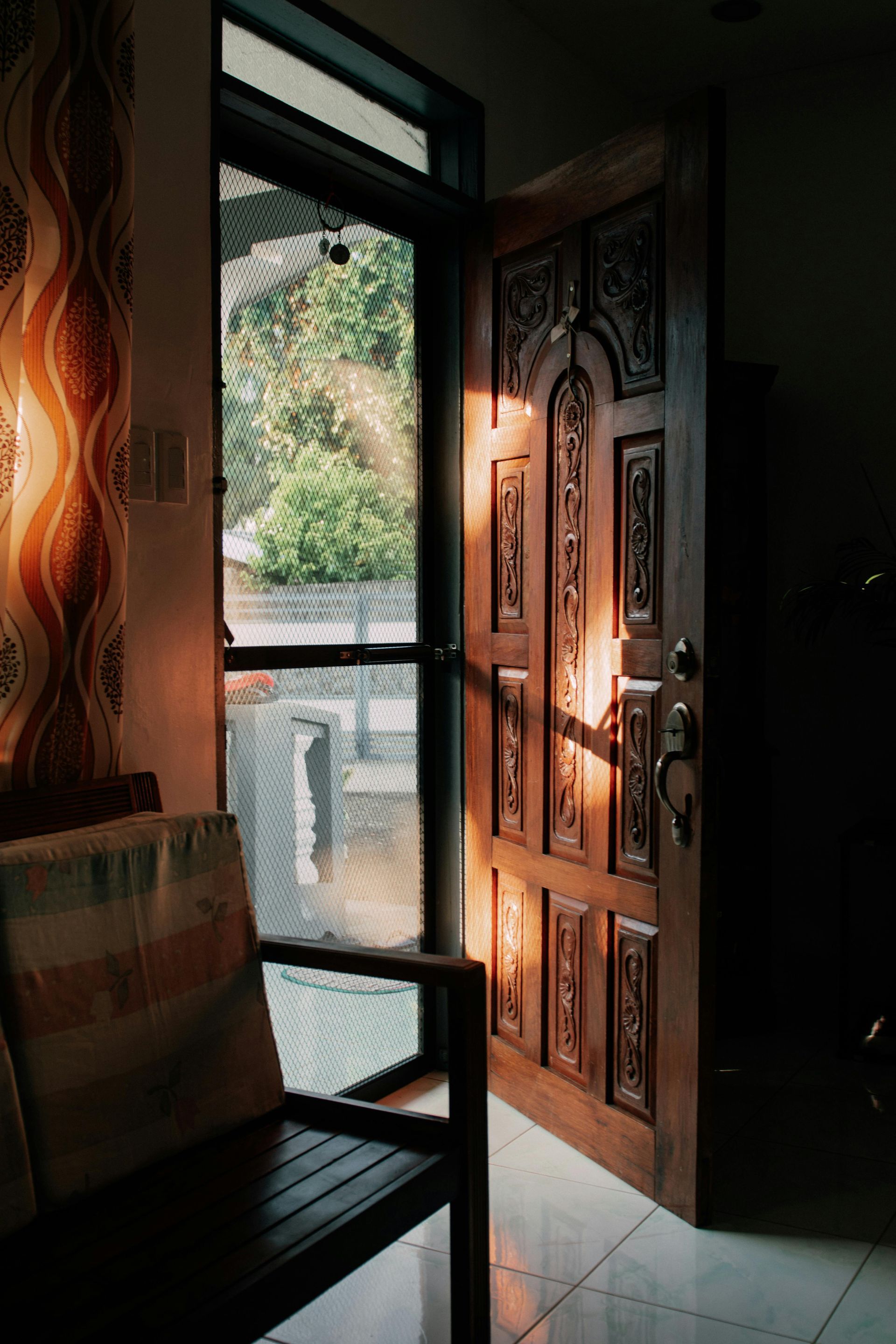 A partially open, carved wooden door is bathed in warm, golden sunlight next to a mesh screen door and a chair.