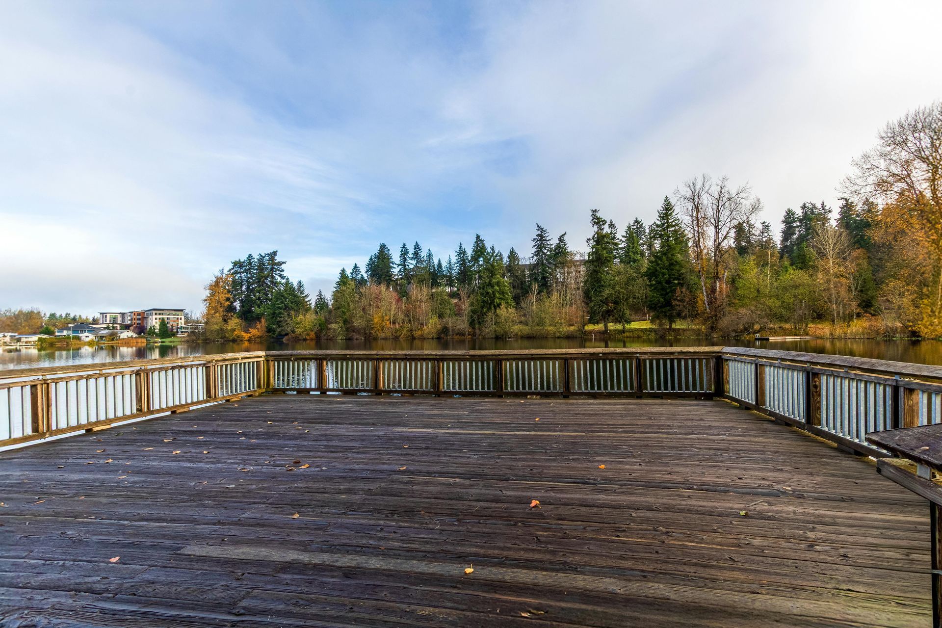 A wooden viewing deck overlooks a calm lake, with trees in autumn colors along the distant shoreline under a cloudy sky.