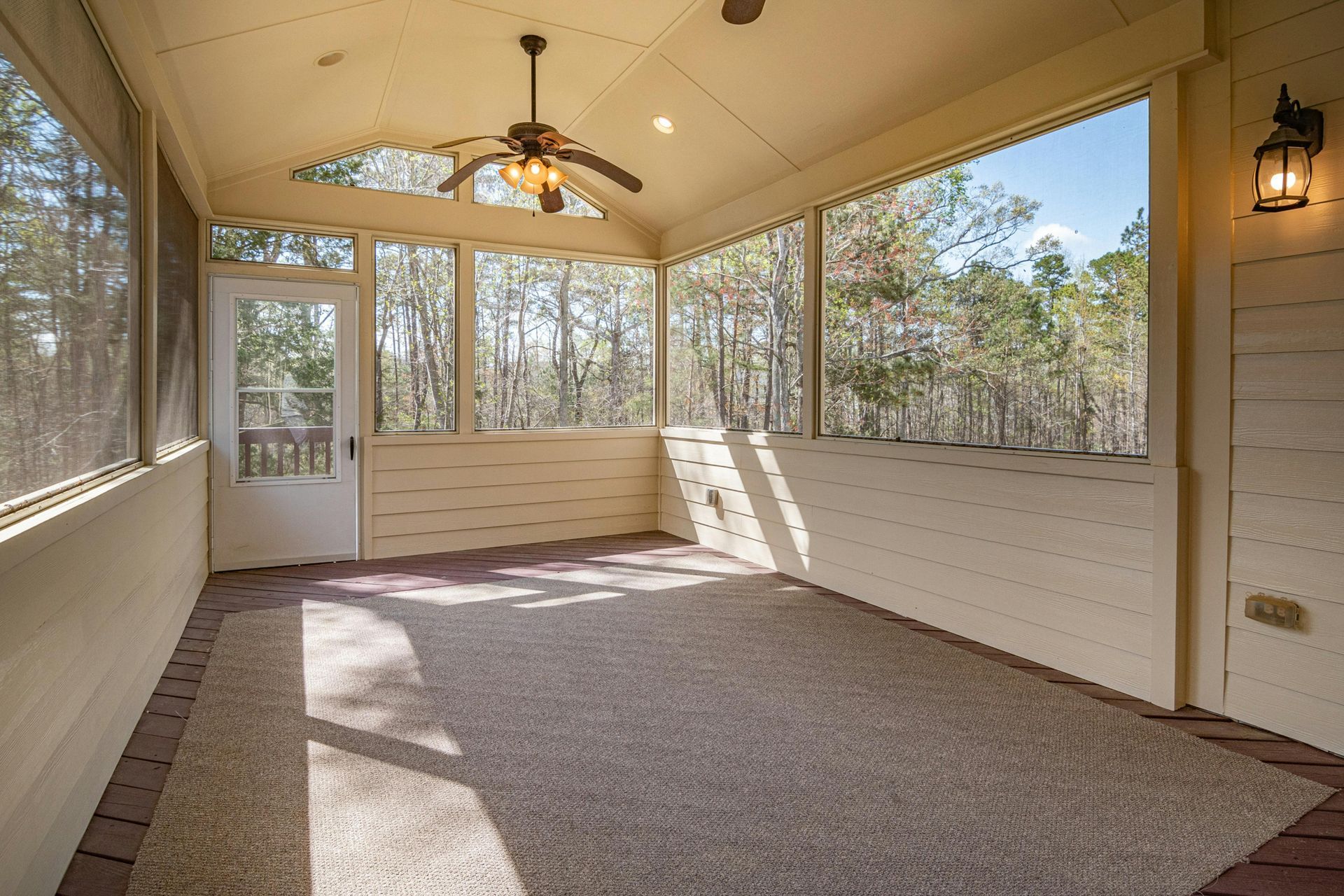 A spacious, empty screened-in porch with light wood paneled walls, a ceiling fan, and large windows overlooking trees.