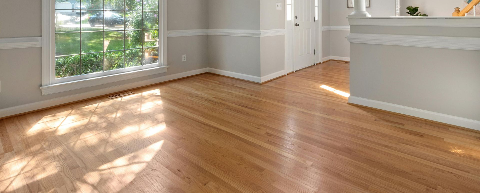 A bright, empty room with polished light wood floors, gray walls, white trim, and a large window.