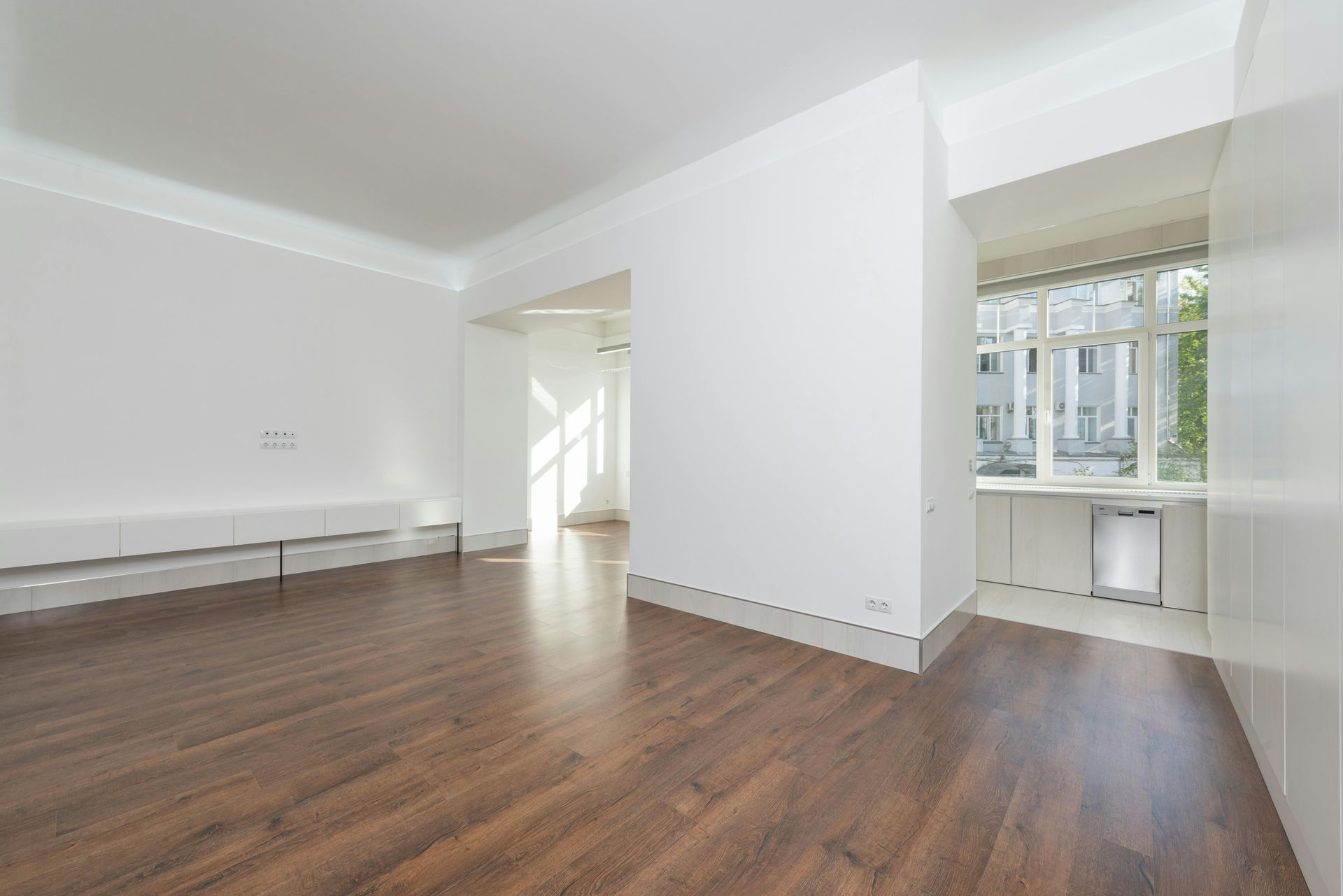 An empty, minimalist living area with white walls, dark wood-look flooring, and a small kitchen nook by a window.