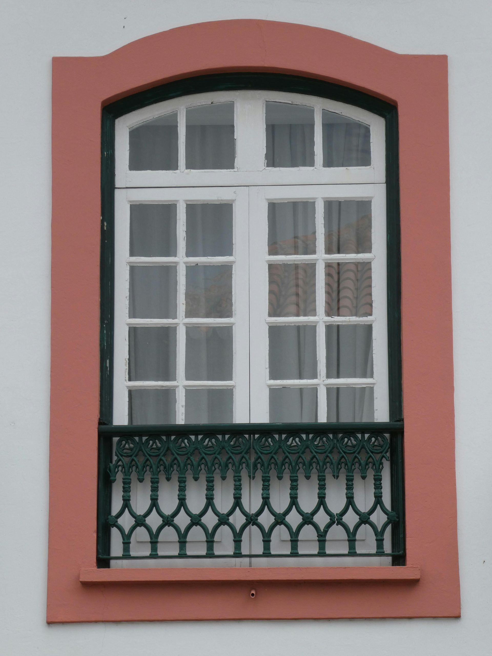 A white-framed window with a small, decorative black iron balcony, set within a light red border on a white wall.