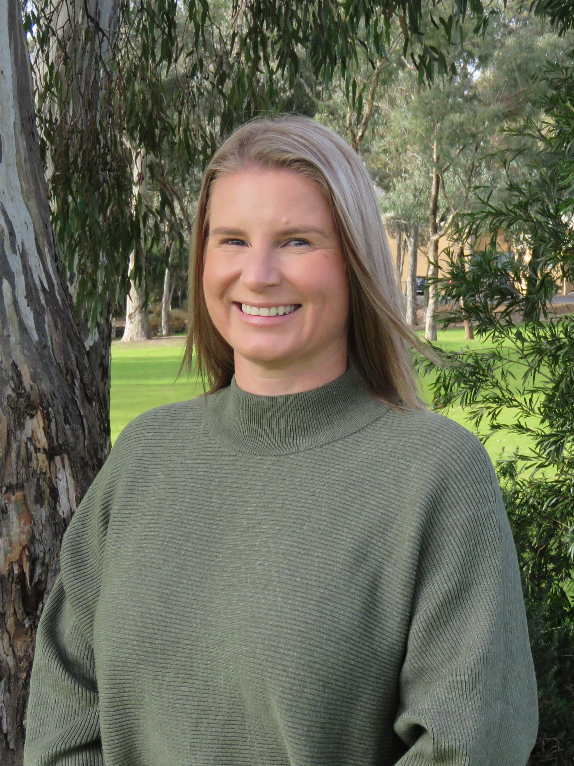 A woman in a green sweater is smiling in front of a tree.