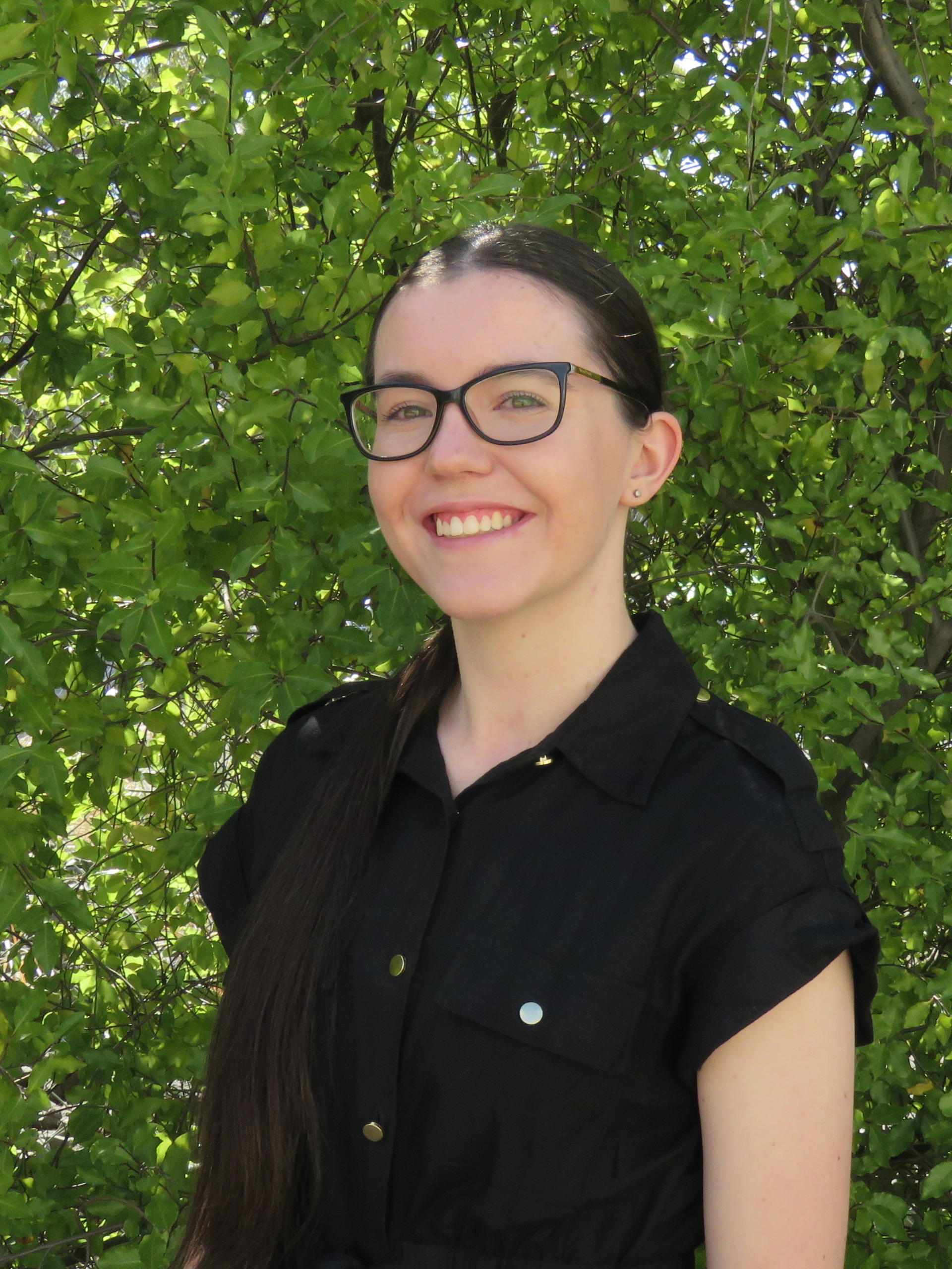 A woman wearing glasses and a black shirt is smiling in front of a tree.