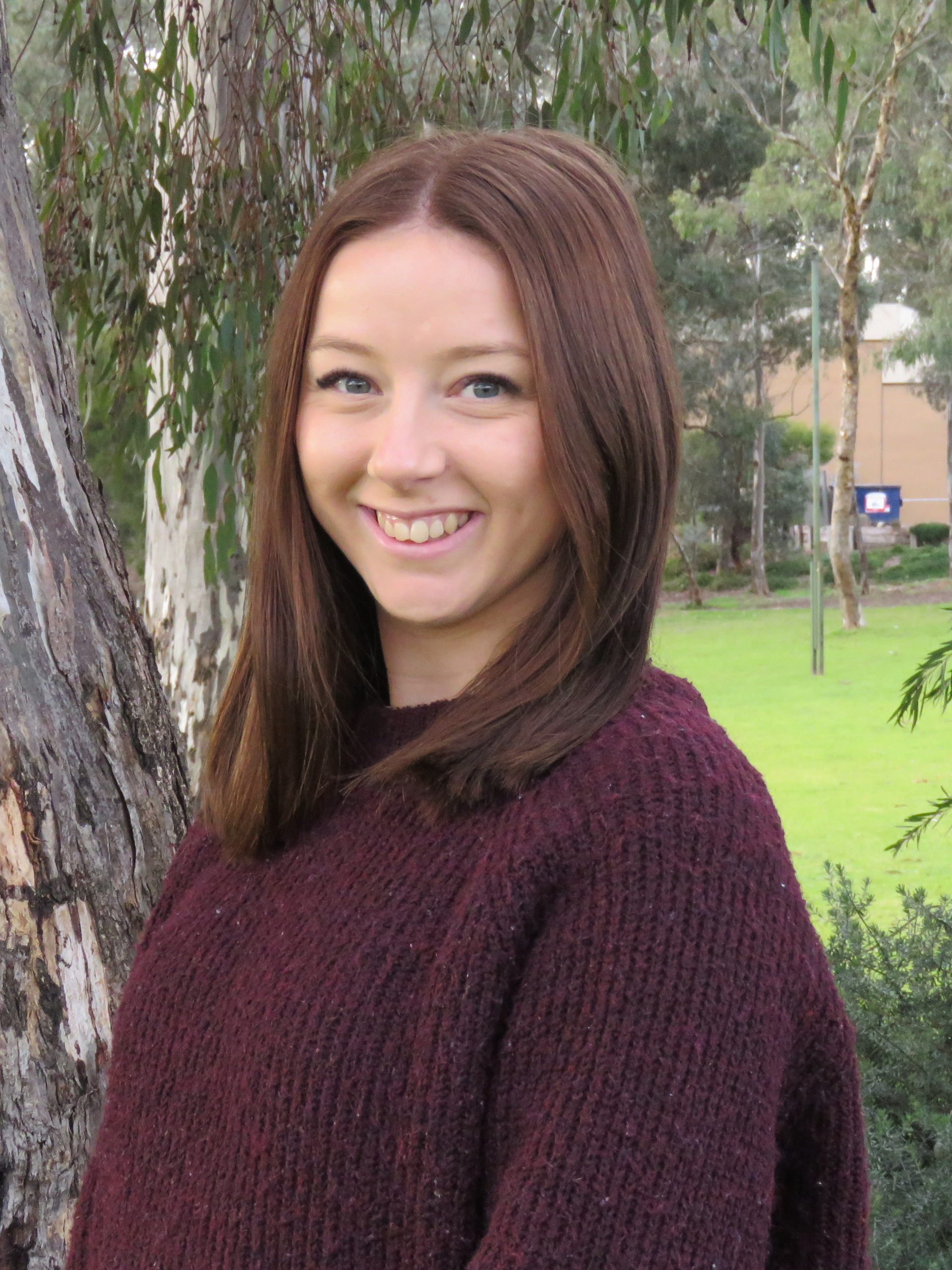 A woman in a maroon sweater is smiling in front of a tree.