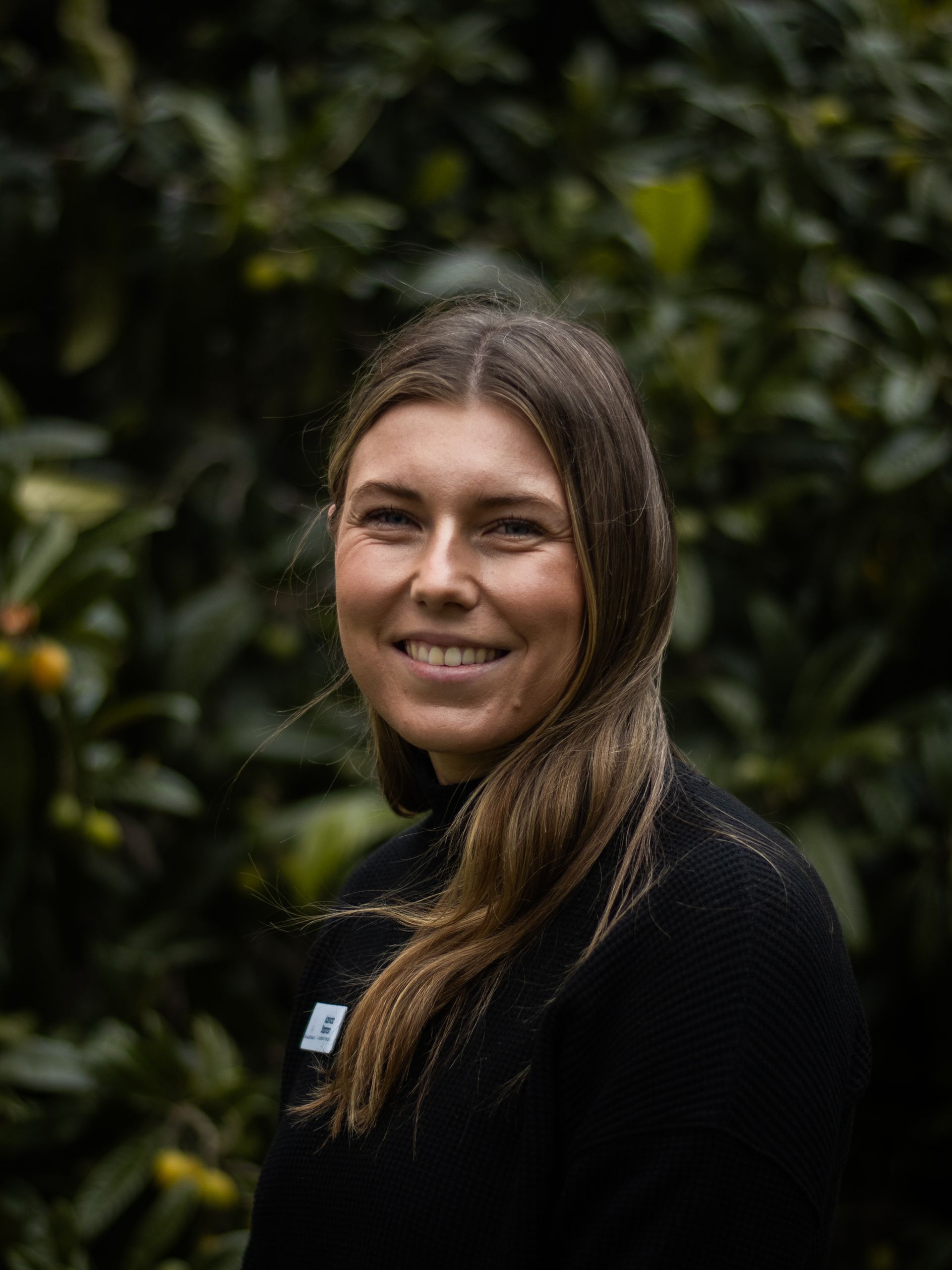 A woman in a black sweater is smiling in front of a bush.