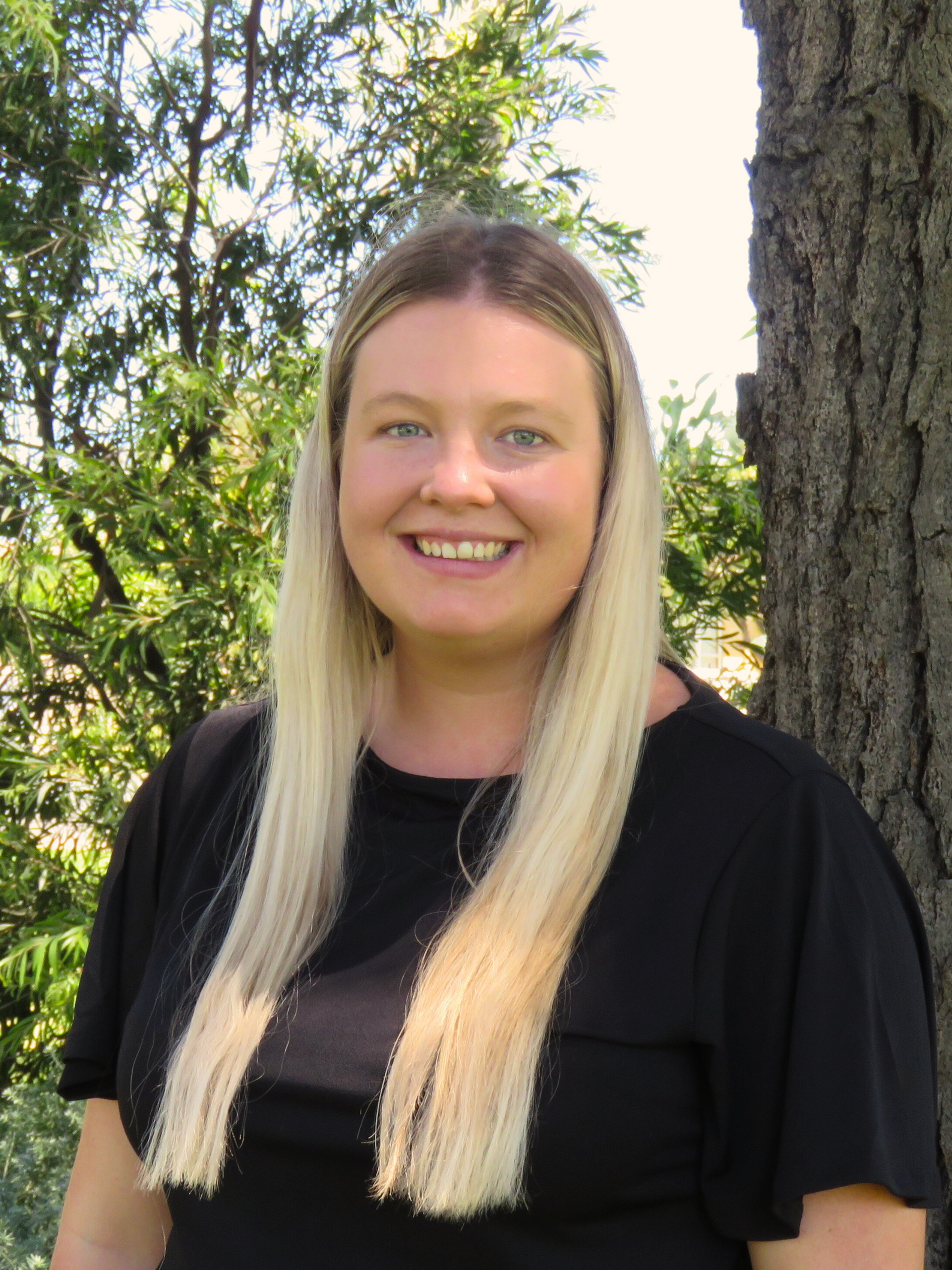 A woman in a black shirt is standing next to a tree and smiling.