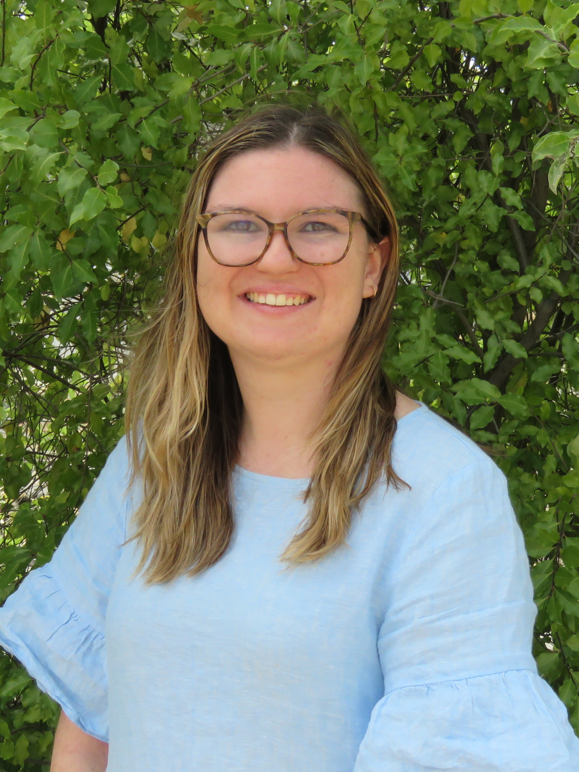 A woman wearing glasses and a blue shirt is smiling in front of a bush.