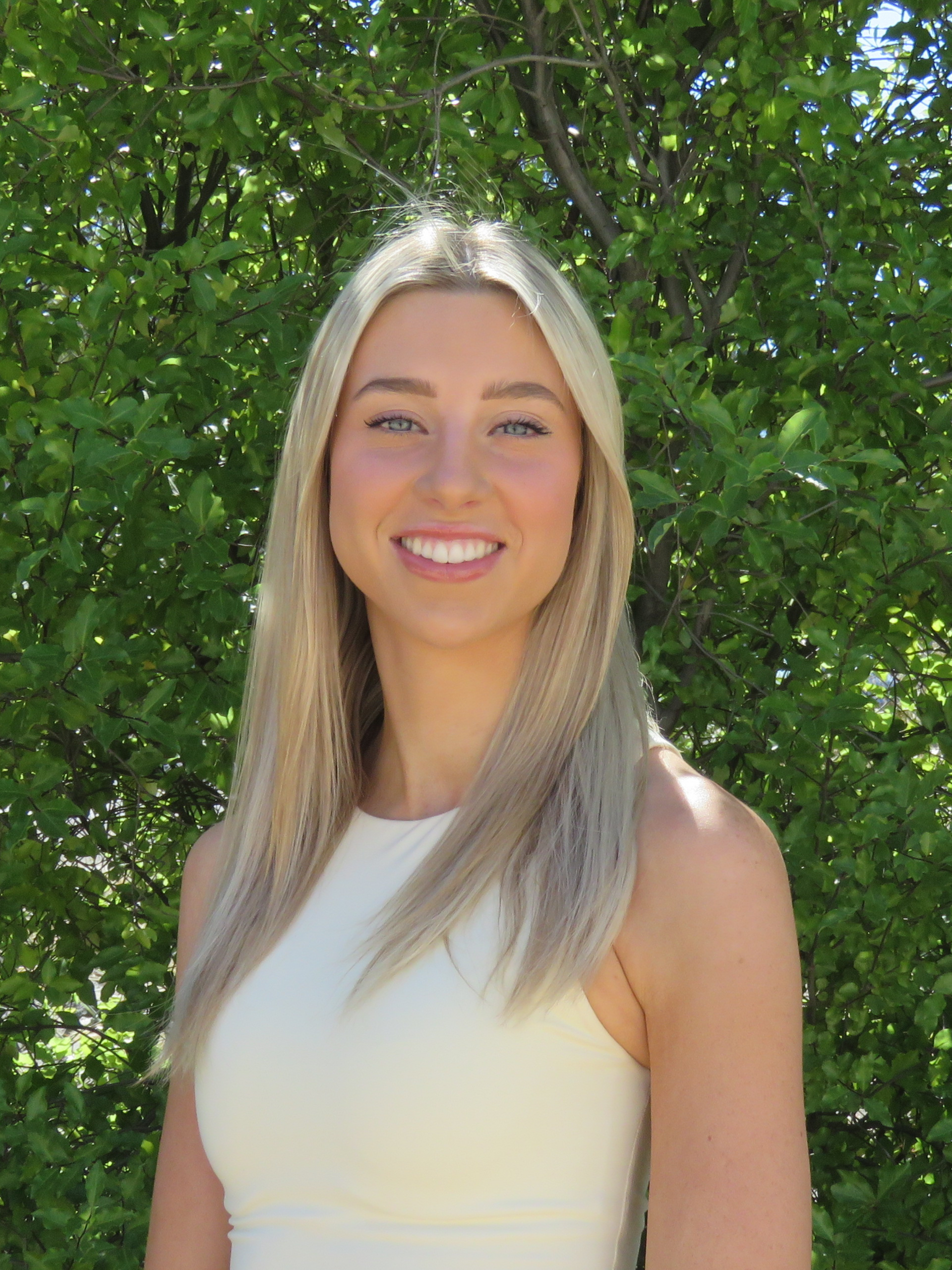 A woman in a white tank top is smiling in front of a tree.