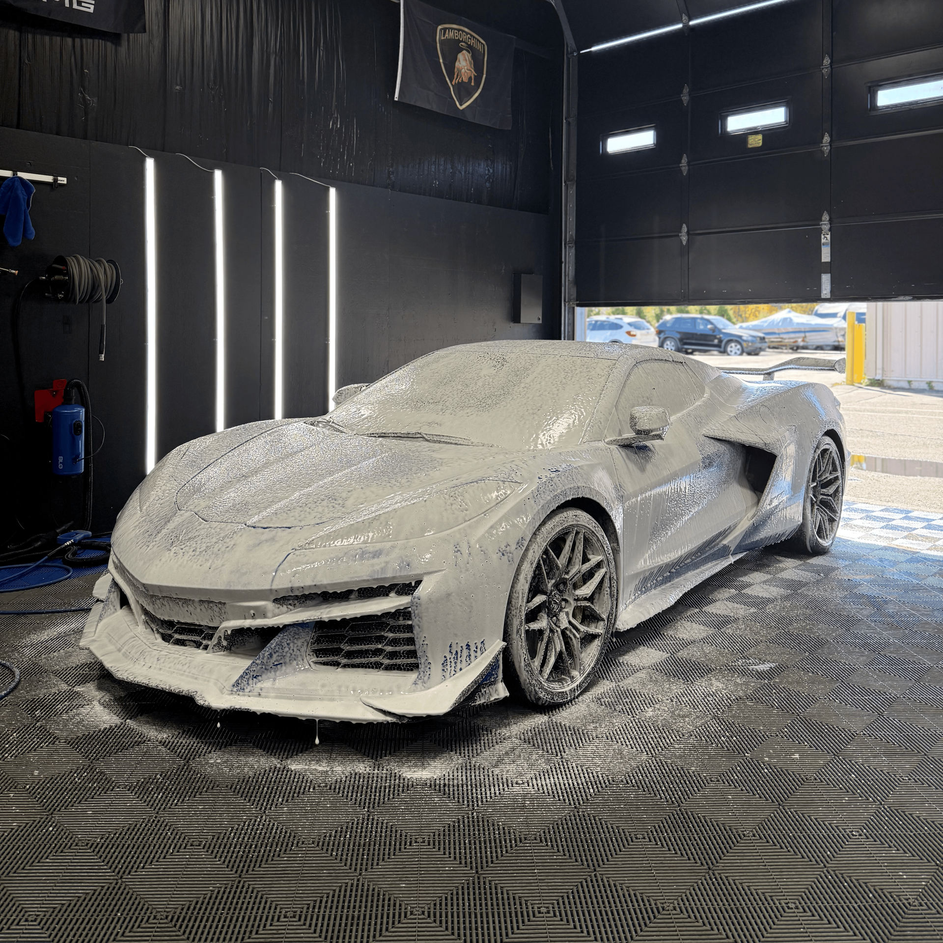 White sports car covered in foam inside a detailing bay, with bright vertical lights and dark walls.