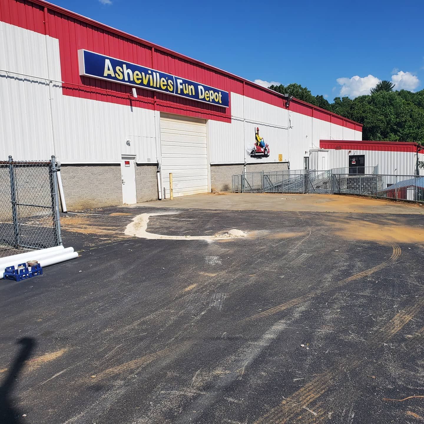 Asheville's Fan Depot building with red and white facade, blue sign, paved parking area, chain-link fence, and sunny sky.