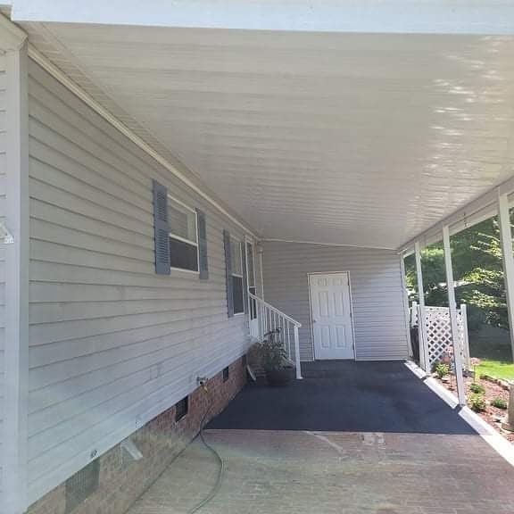 Covered carport attached to a light gray house with blue shutters, a white door, and black asphalt.