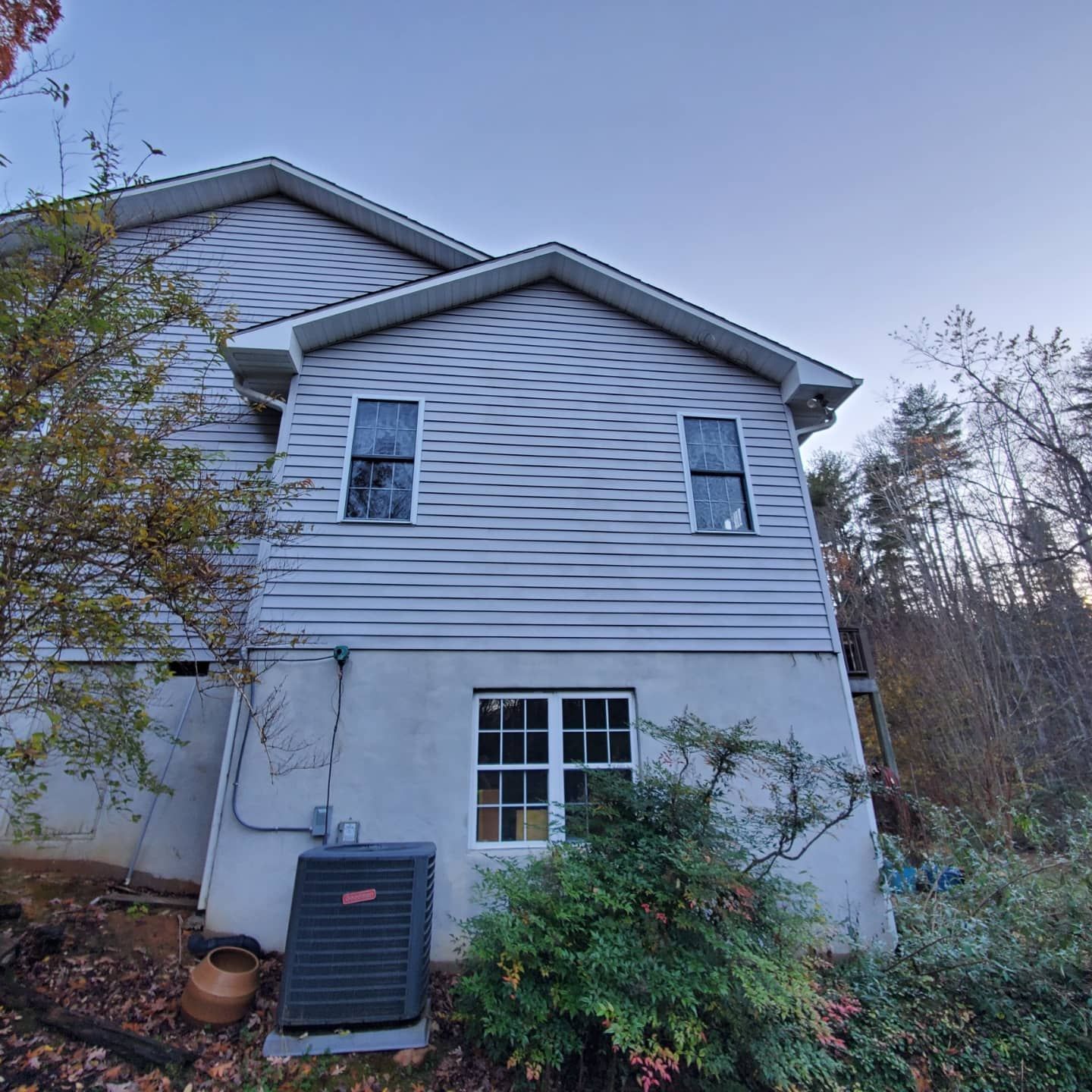 Side view of a two-story light gray house with windows, air conditioning unit, and foliage in a wooded setting.