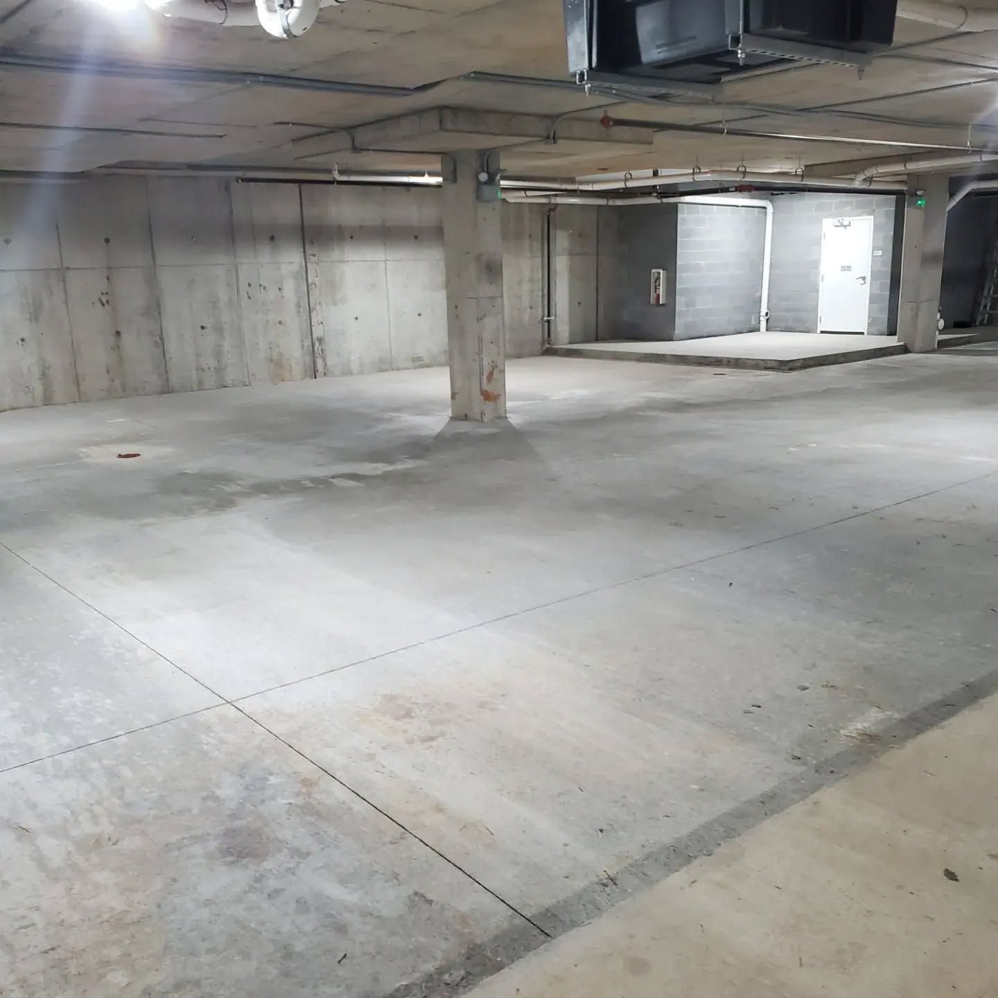 Concrete parking garage with pillars, walls, and a door in the background. Overhead lights and pipes.