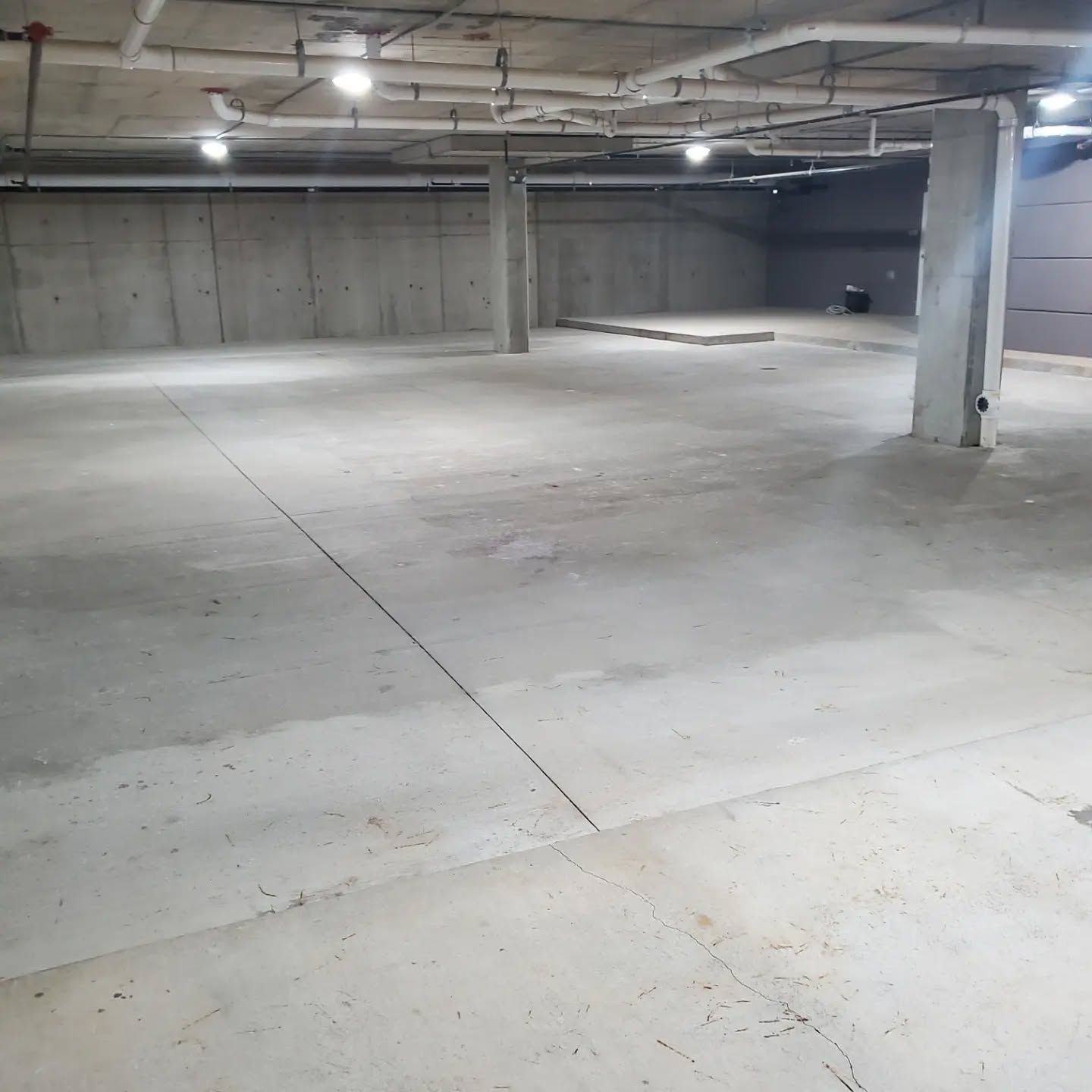 Empty, unfinished concrete interior with support pillars, overhead pipes, and lighting.