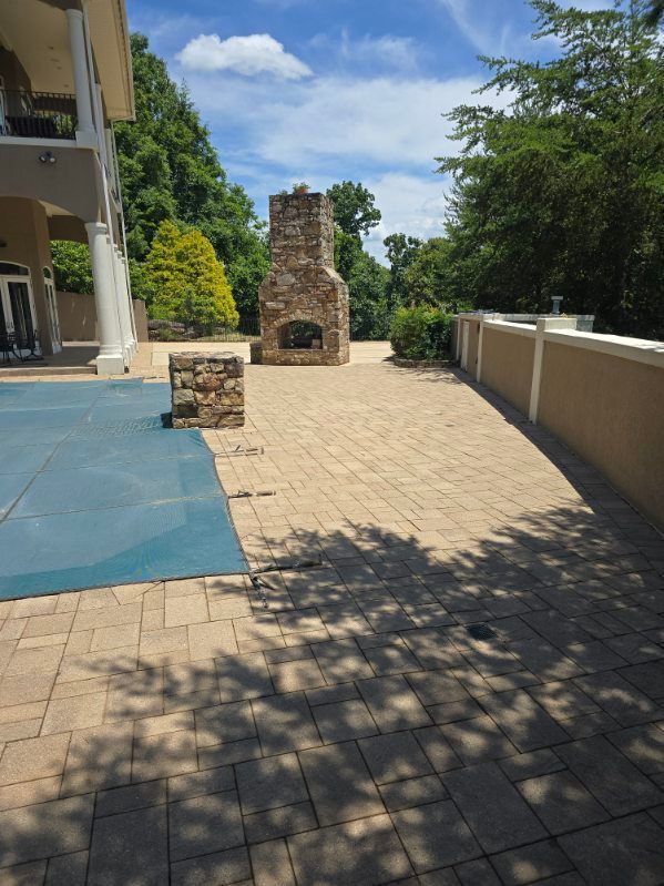 Stone patio with an outdoor fireplace, near a pool and a building, under a partly cloudy sky.
