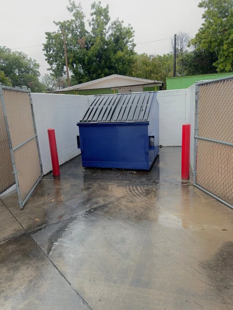 Blue dumpster enclosed by chain-link fences and concrete in front of a white wall, under an overcast sky.