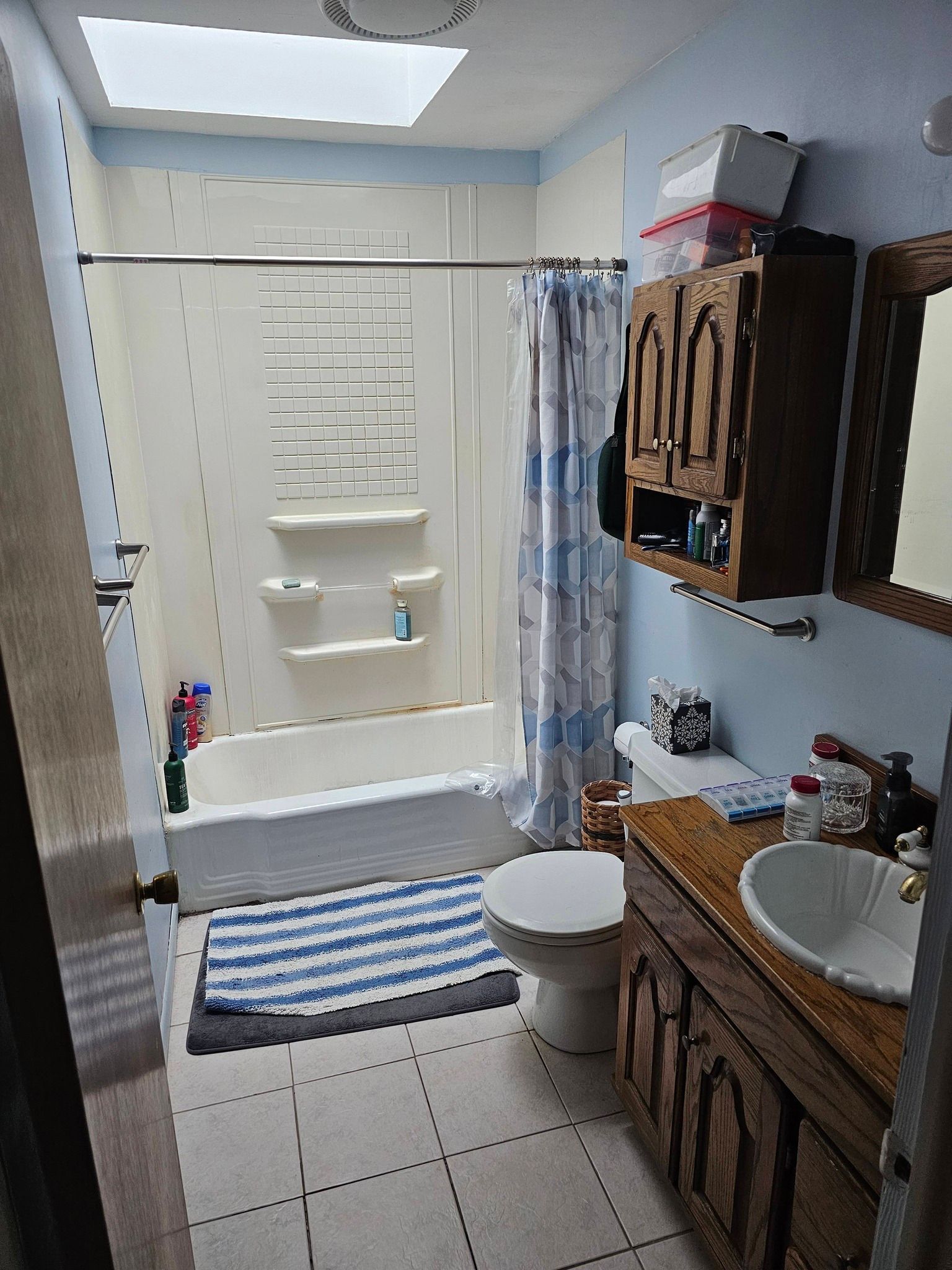 A bathroom with a bathtub shower combo, blue and white striped mat, white toilet, and a wooden vanity with a white sink.