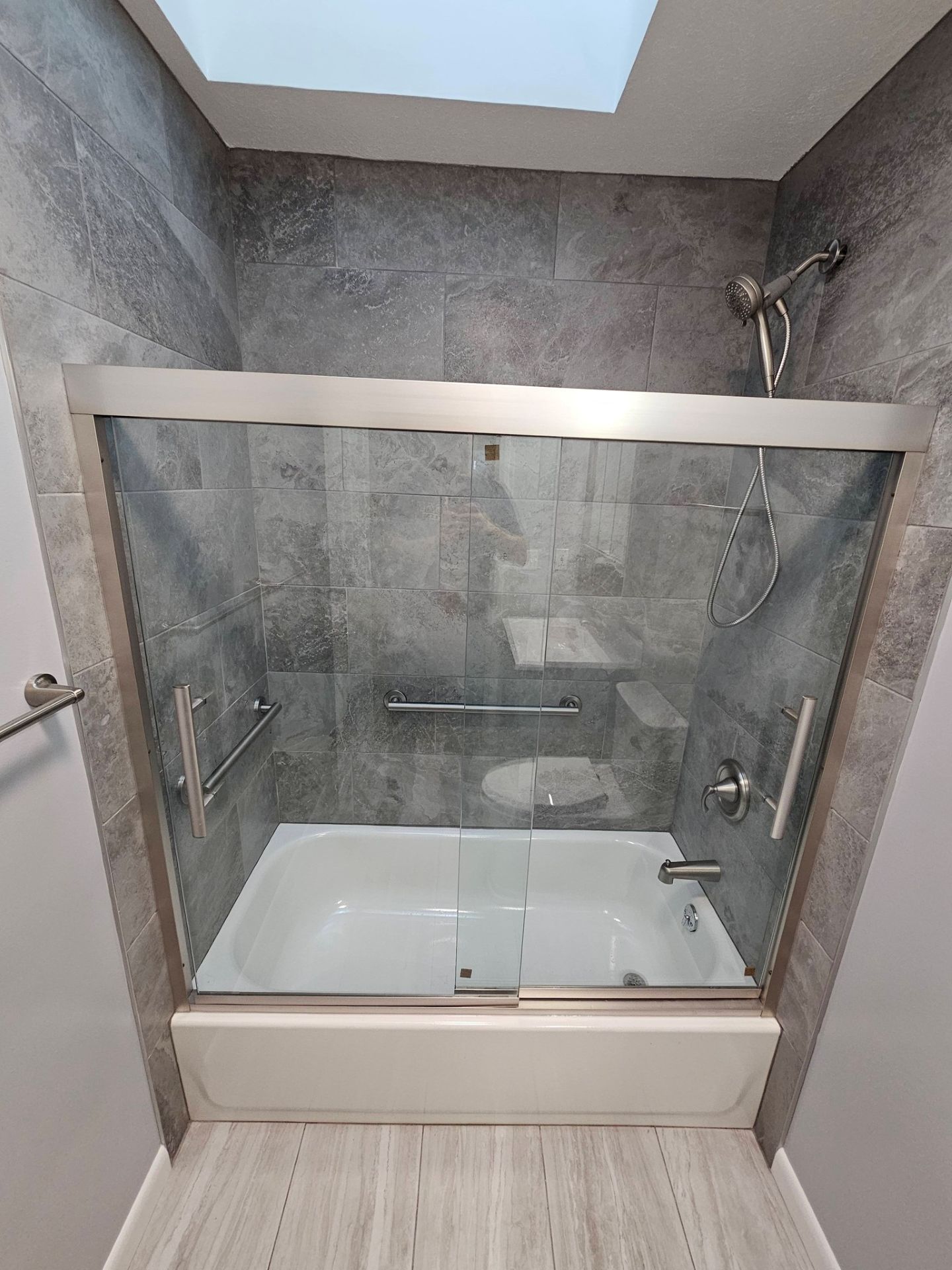 A bathroom with a white bathtub, glass sliding doors, and a shower head against a grey tiled wall under a skylight.