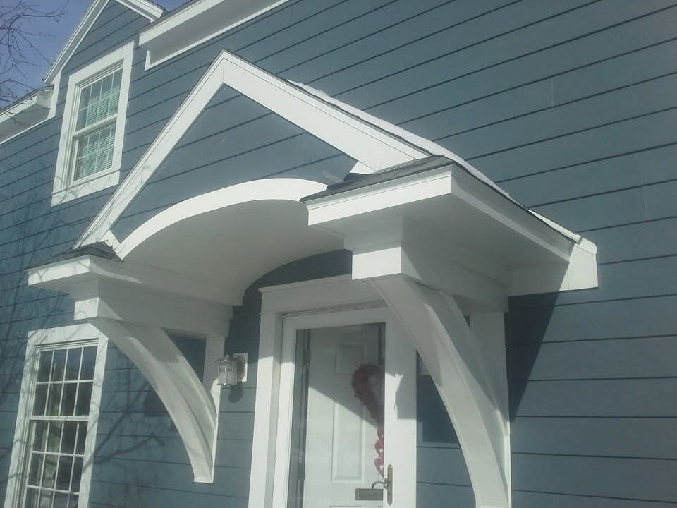 A white gabled door hood with curved decorative brackets attached to a dark blue, wood-sided house.