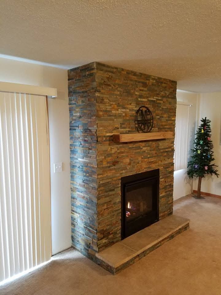 A fireplace with a stacked stone facade and wooden mantel, situated next to a sliding glass door and a small Christmas tree.