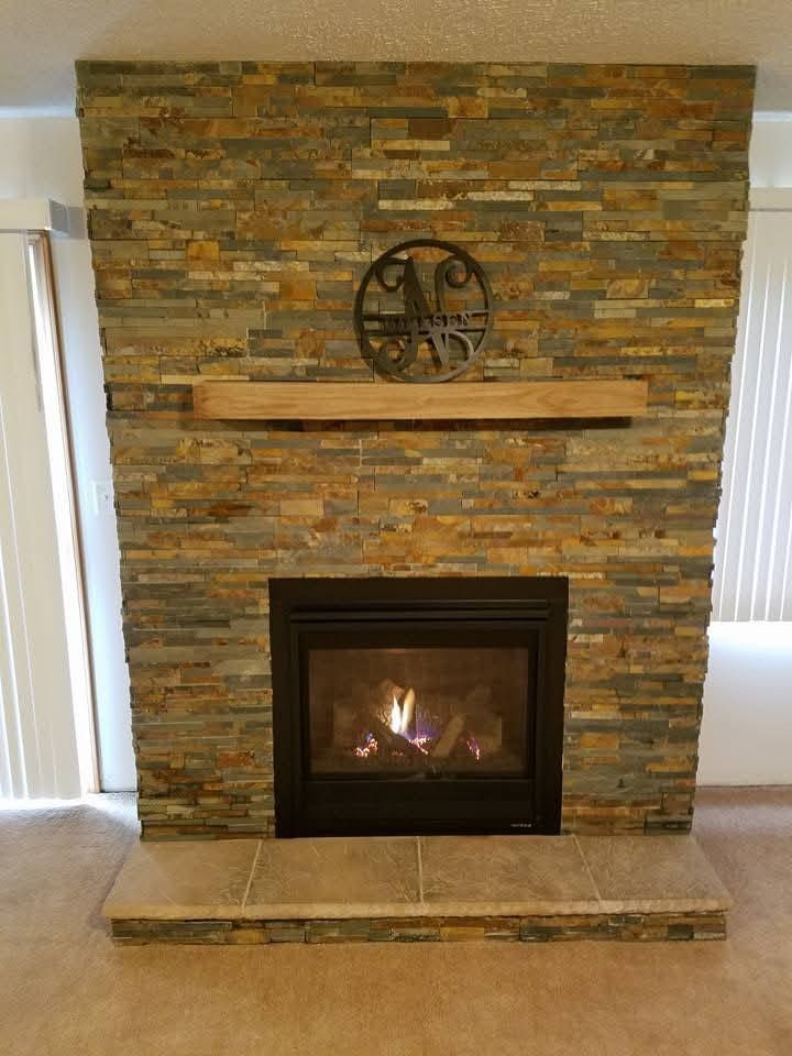 A fireplace with a lit fire, stone-textured surround, wooden mantel, and a decorative circular metal initial on the wall.