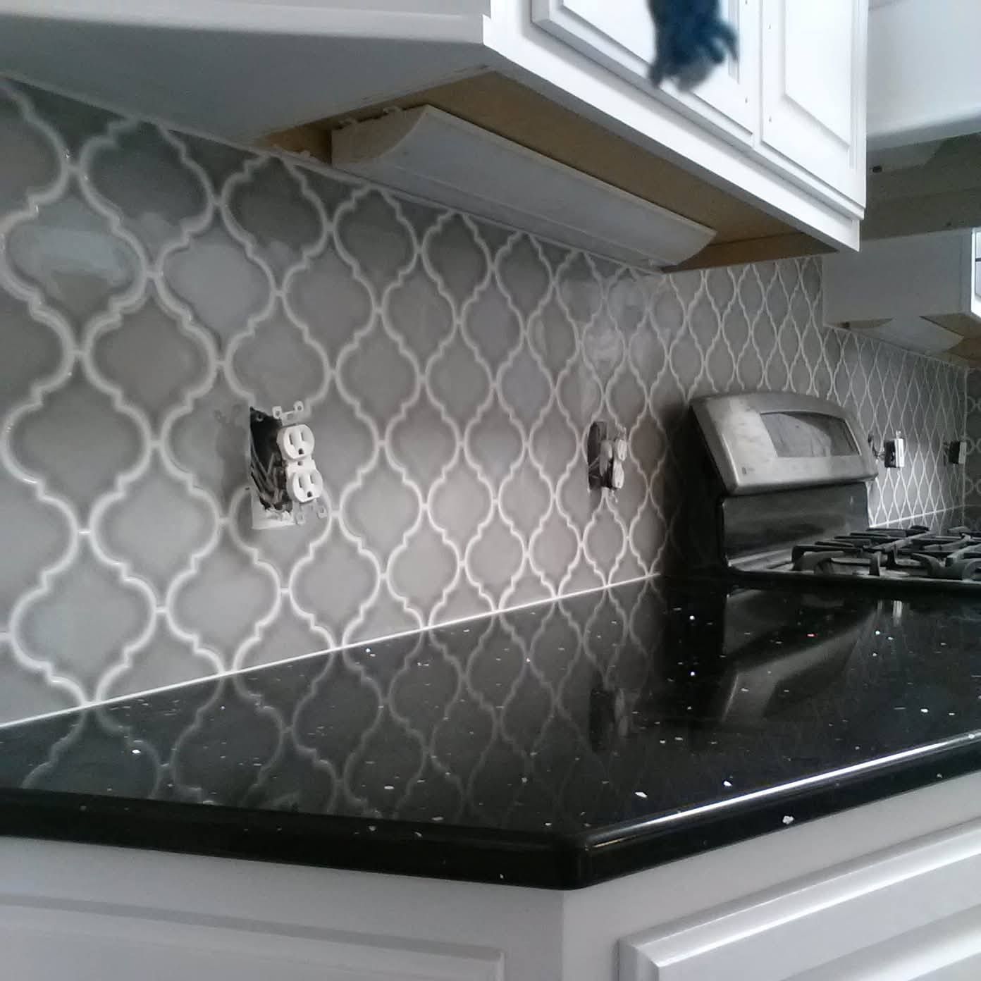 A kitchen backsplash with grey, Moroccan-style tiles and white grout, set above a black countertop and white cabinets.