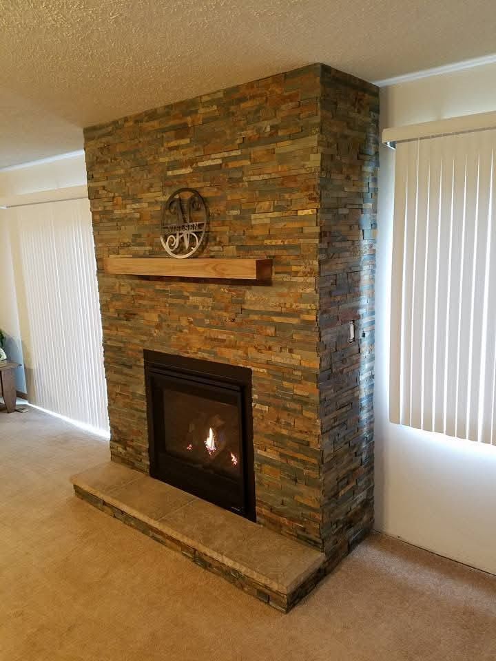 A fireplace with a stacked stone facade, a thick wooden mantel, a large stone hearth, and a glass-enclosed gas fire.