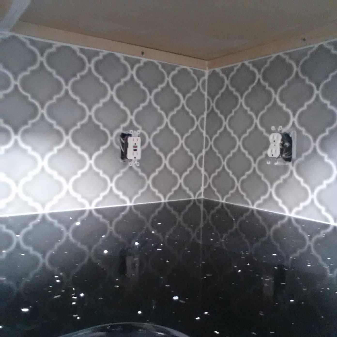 A kitchen backsplash featuring gray and white arabesque tiles, a black speckled countertop, and two electrical outlets.
