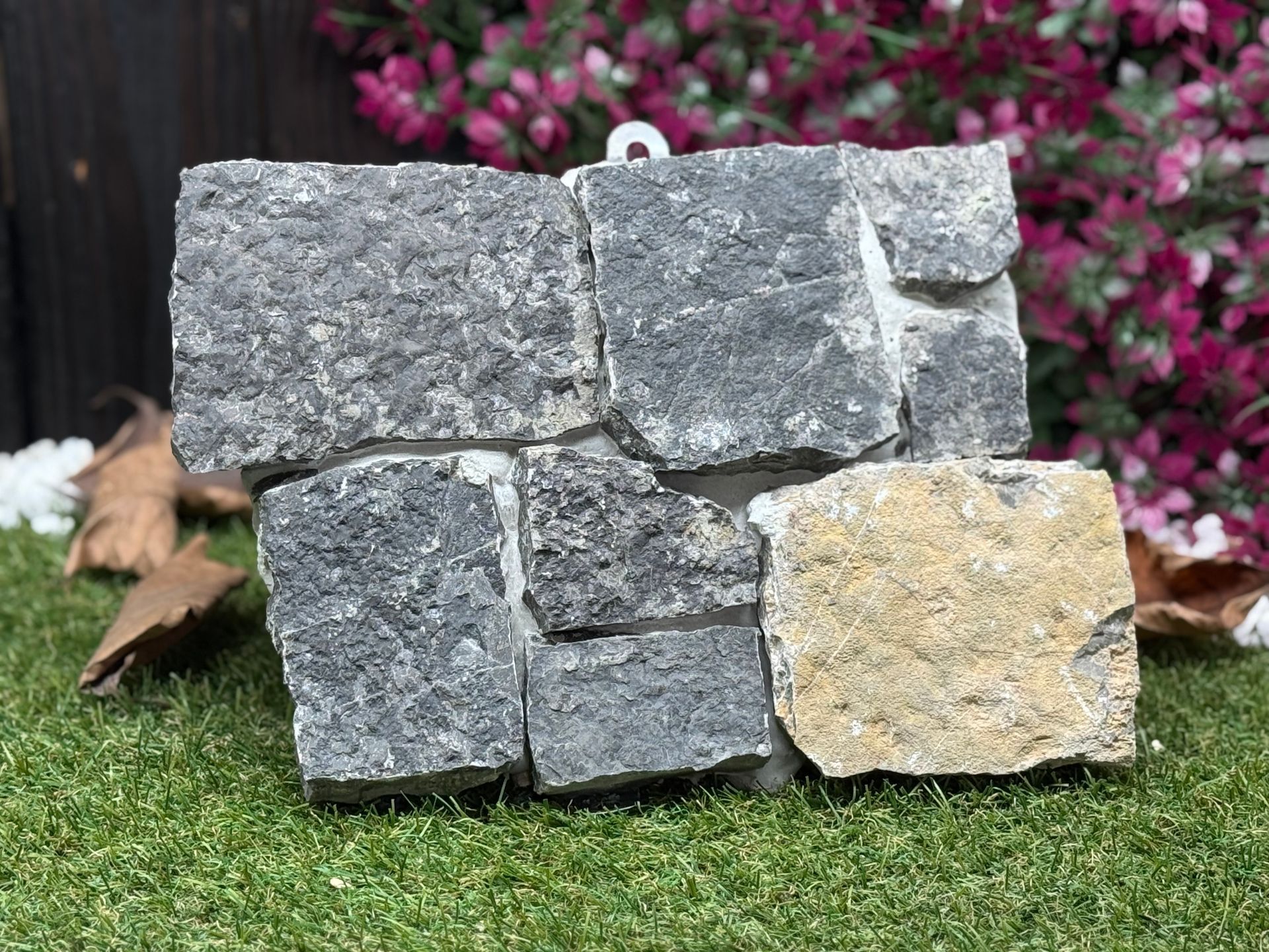 Eine kleine, dekorative Steinmauer aus grauen und hellbraunen Steinen auf grünem Gras vor einem floralen Hintergrund.
