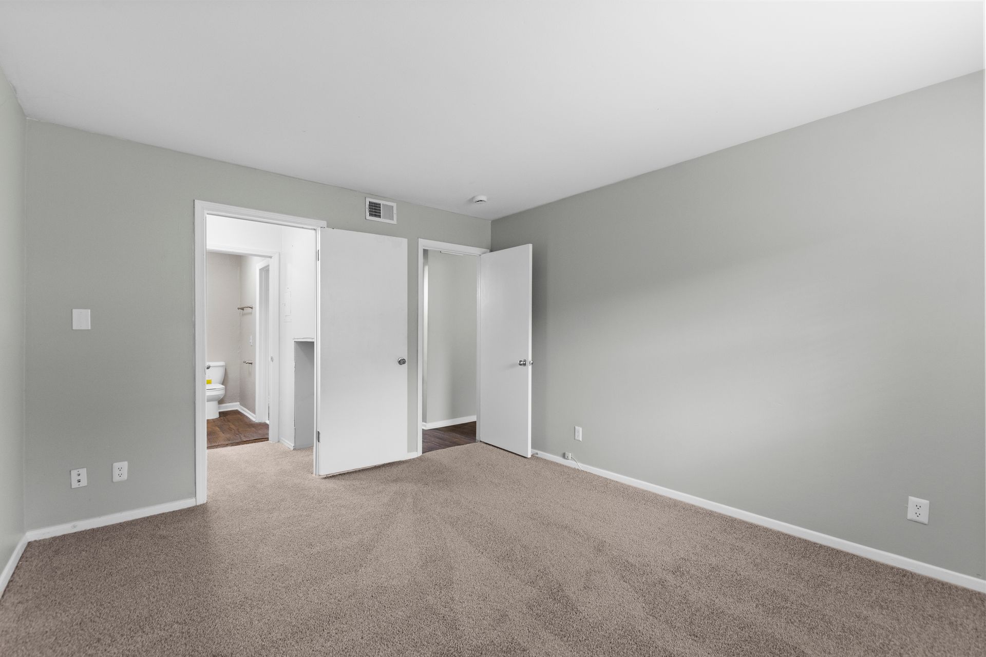 An empty bedroom with gray walls, beige carpet, and two open white doors leading to an adjacent room.