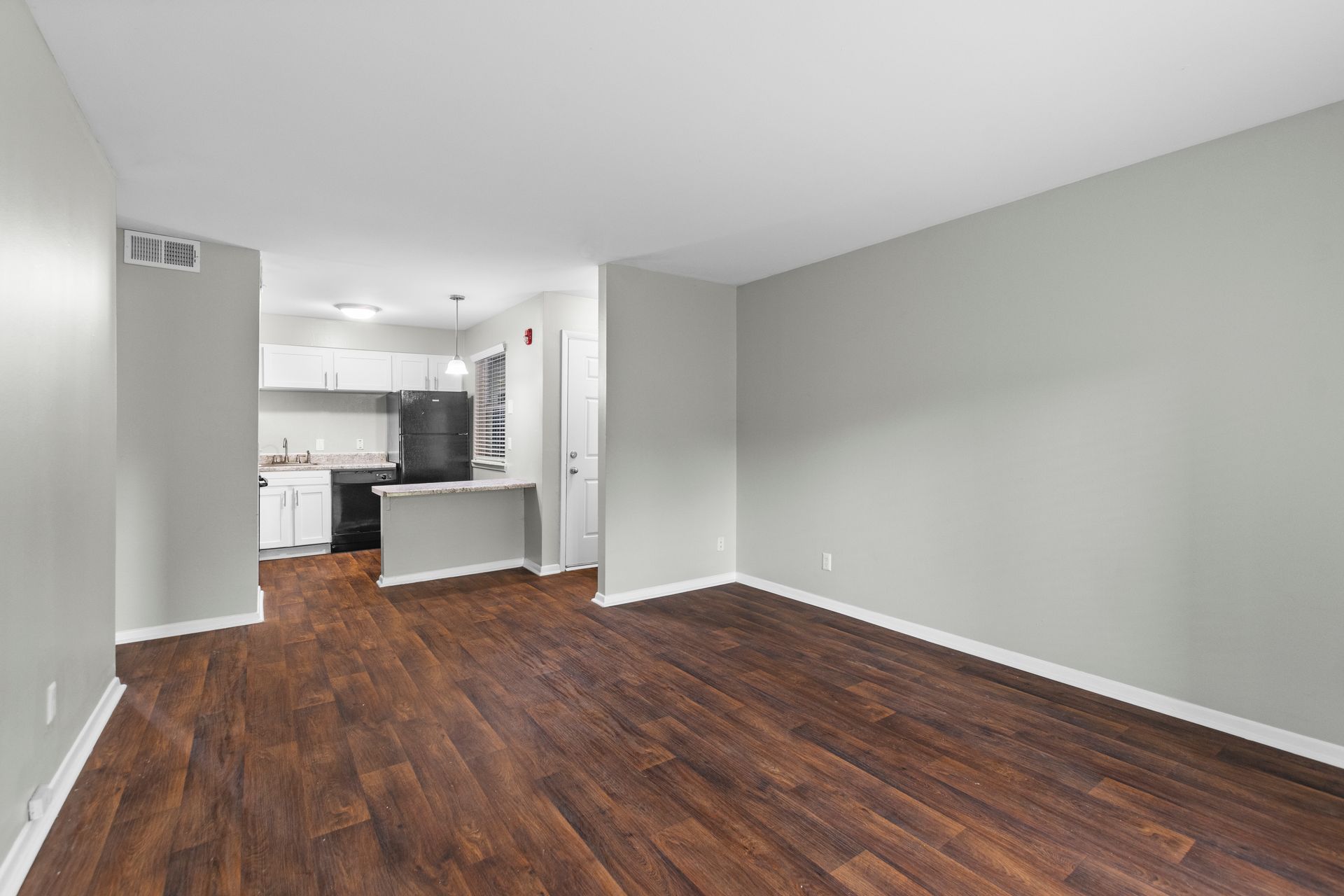 An empty living room with wood-look flooring and light gray walls, leading to a small kitchen with dark appliances.