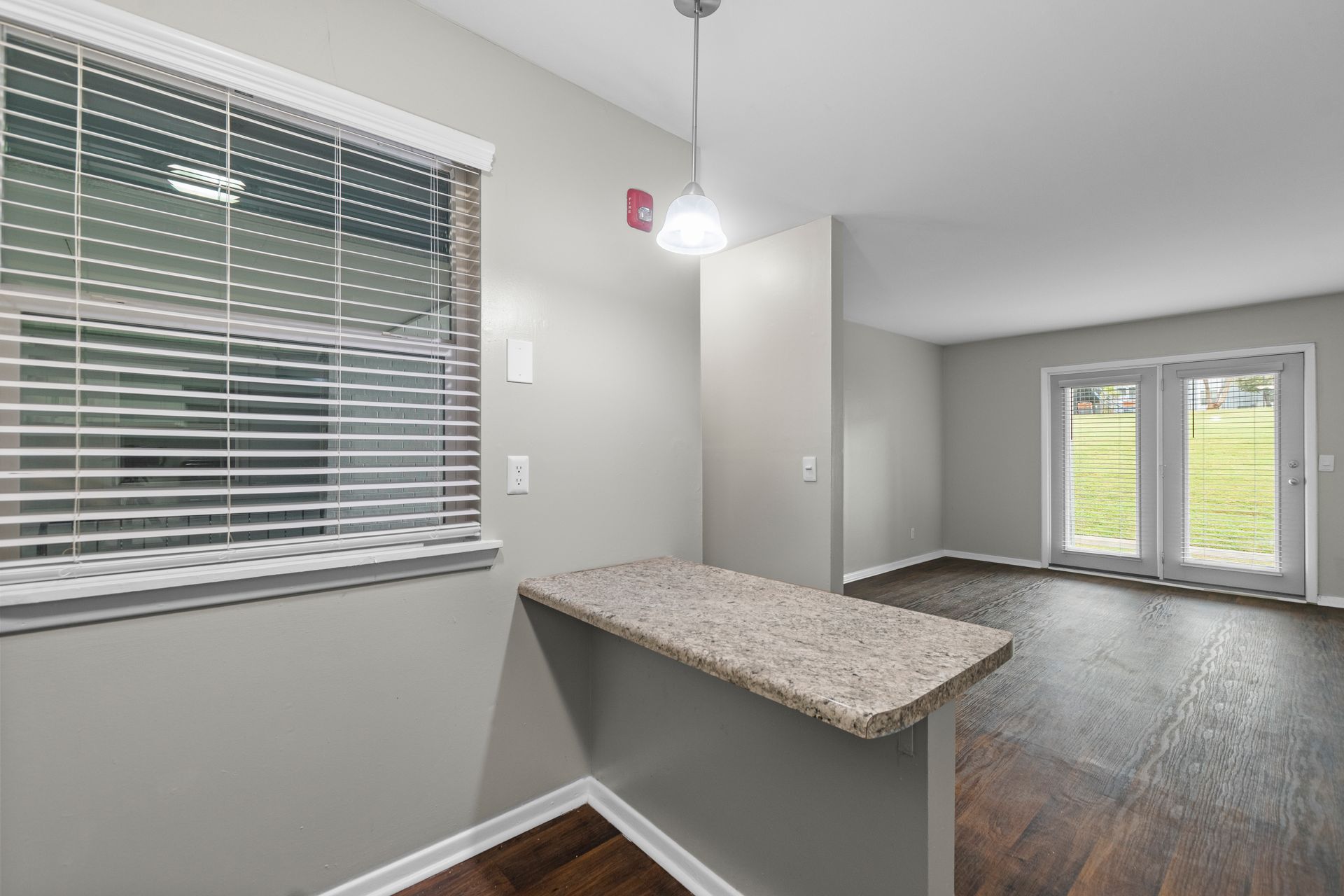 A grey kitchen area with a granite bar counter, a window with horizontal blinds, and a view of glass doors to a yard.